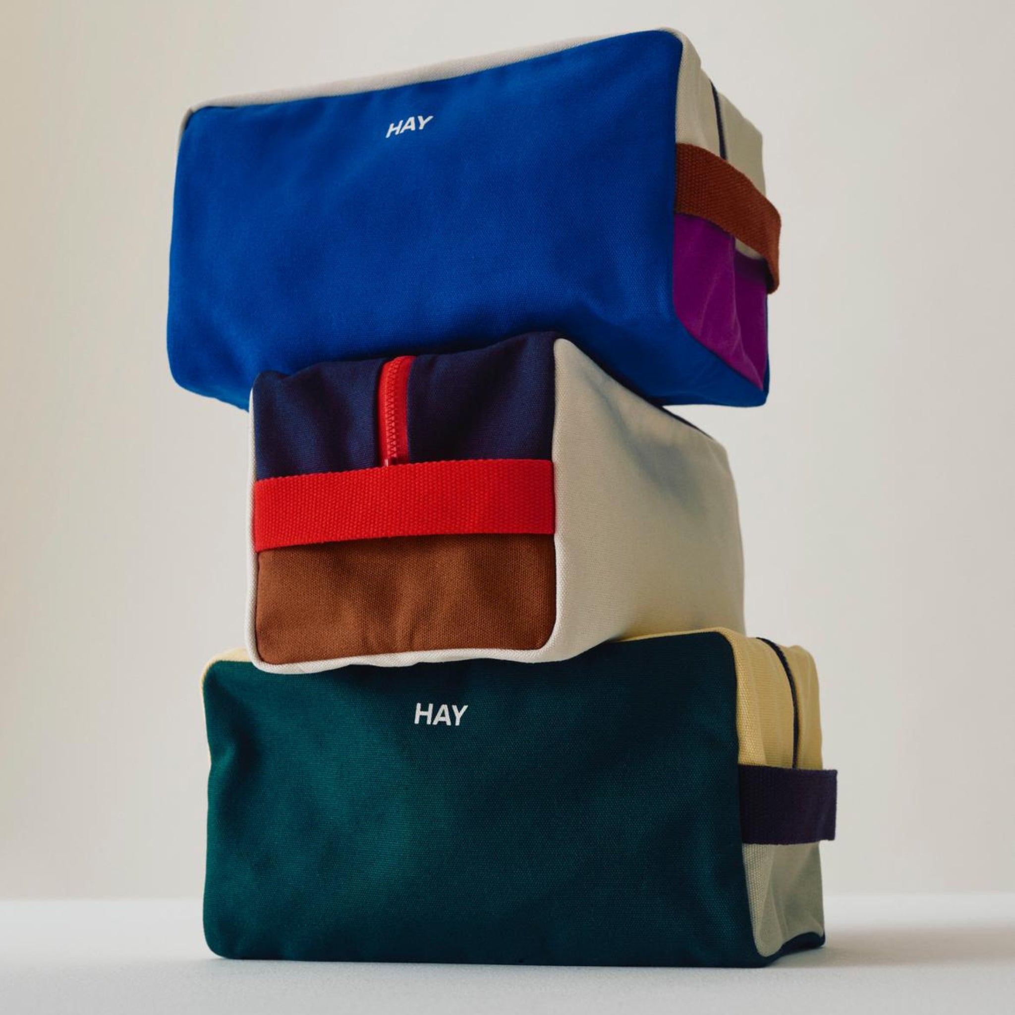 Drie rechthoekige HAY Multi Toilettas Medium Crème zijn gestapeld, elk gemaakt van biologisch katoen met een bijpassend handvat en rits. De merknaam "HAY" staat op de voorkant. De achtergrond is effen en licht.