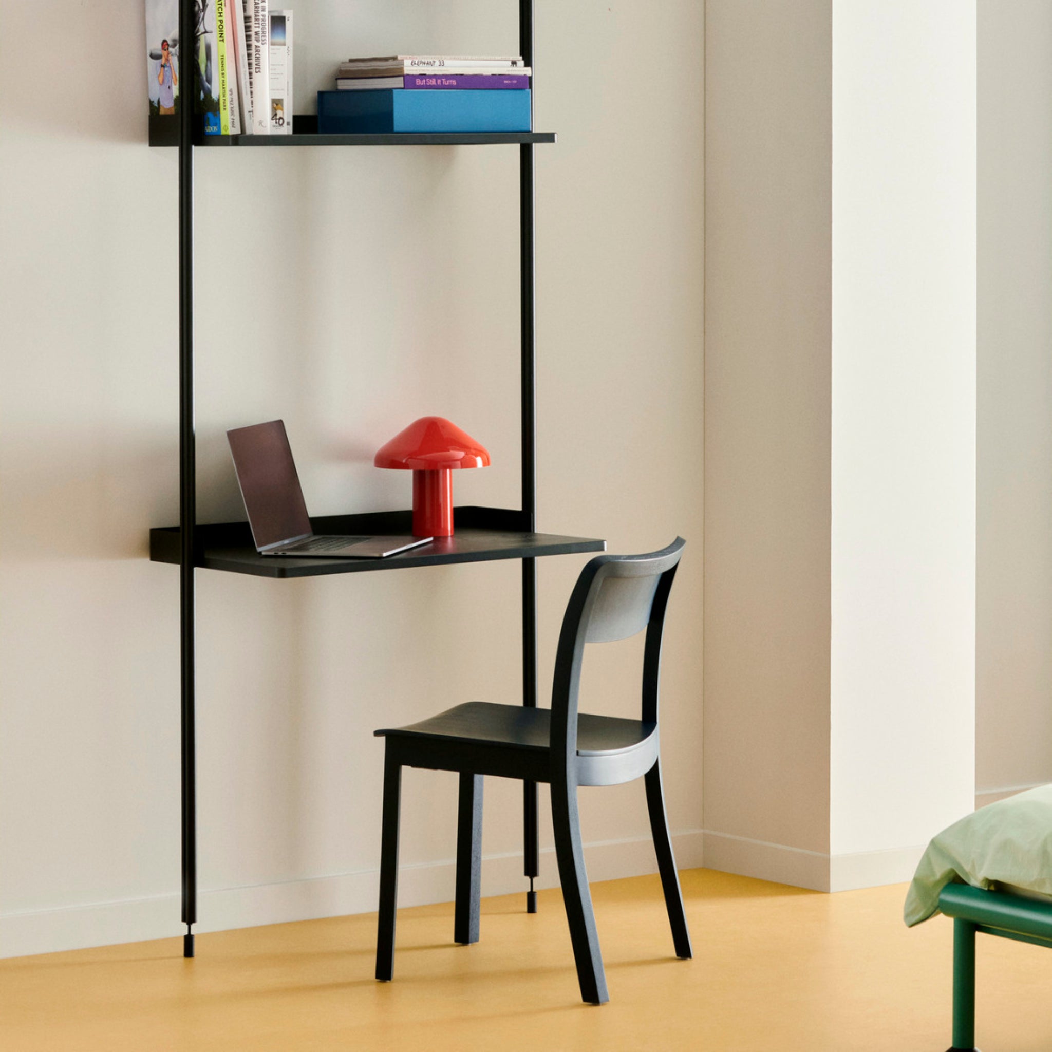Een minimalistische werkplek met een zwart zwevend bureau, grijze stoel, open laptop en een kleine rode Hay Pao Tafellamp LED Oplaadbaar Rood van Hay. Boven het bureau is een plank met boeken, dit alles tegen een lichte crèmekleurige muur en gele vloer.