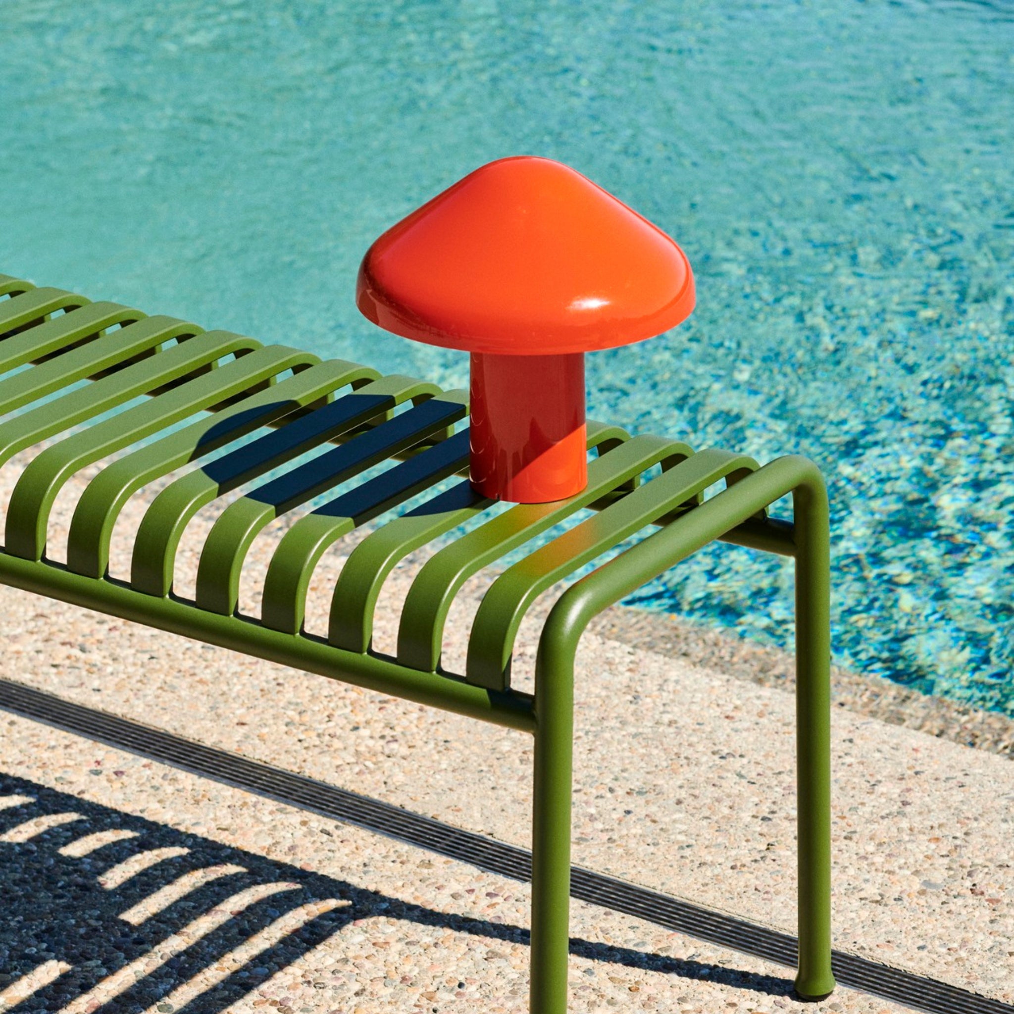 Een groene metalen lattenbank staat naast een zwembad, met daarbovenop de Hay Pao Tafellamp LED Oplaadbaar Rood van Hay, een opvallende rode draadloze lamp, geplaatst aan de rand van het zwembad.