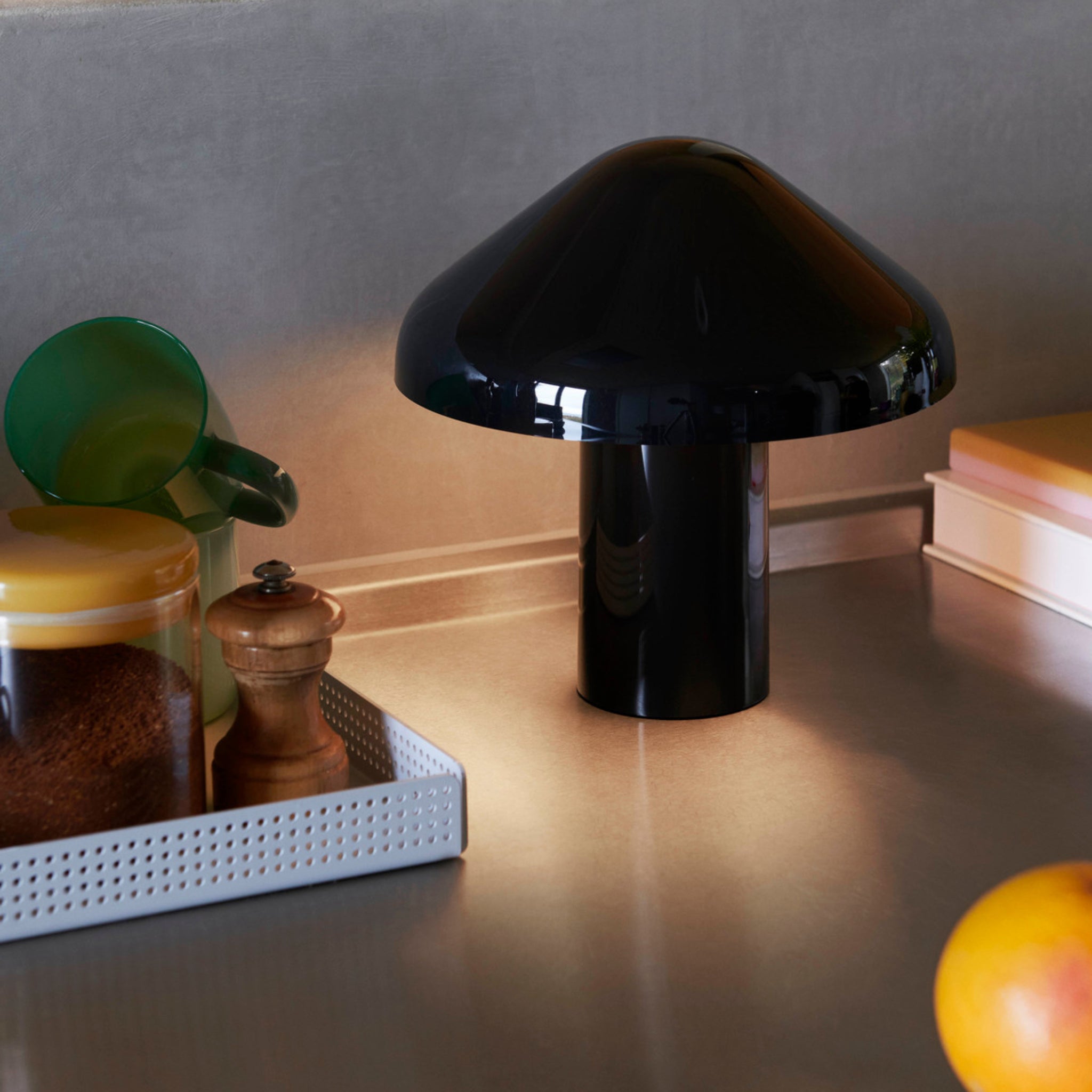 Een Hay Pao Tafellamp LED Oplaadbaar Zwart van Hay, ontworpen door Naoto Fukasawa, staat op een aanrecht naast een groen kopje, potje met geel deksel, pepermolen, boeken en een deels zichtbare sinaasappel op de voorgrond.