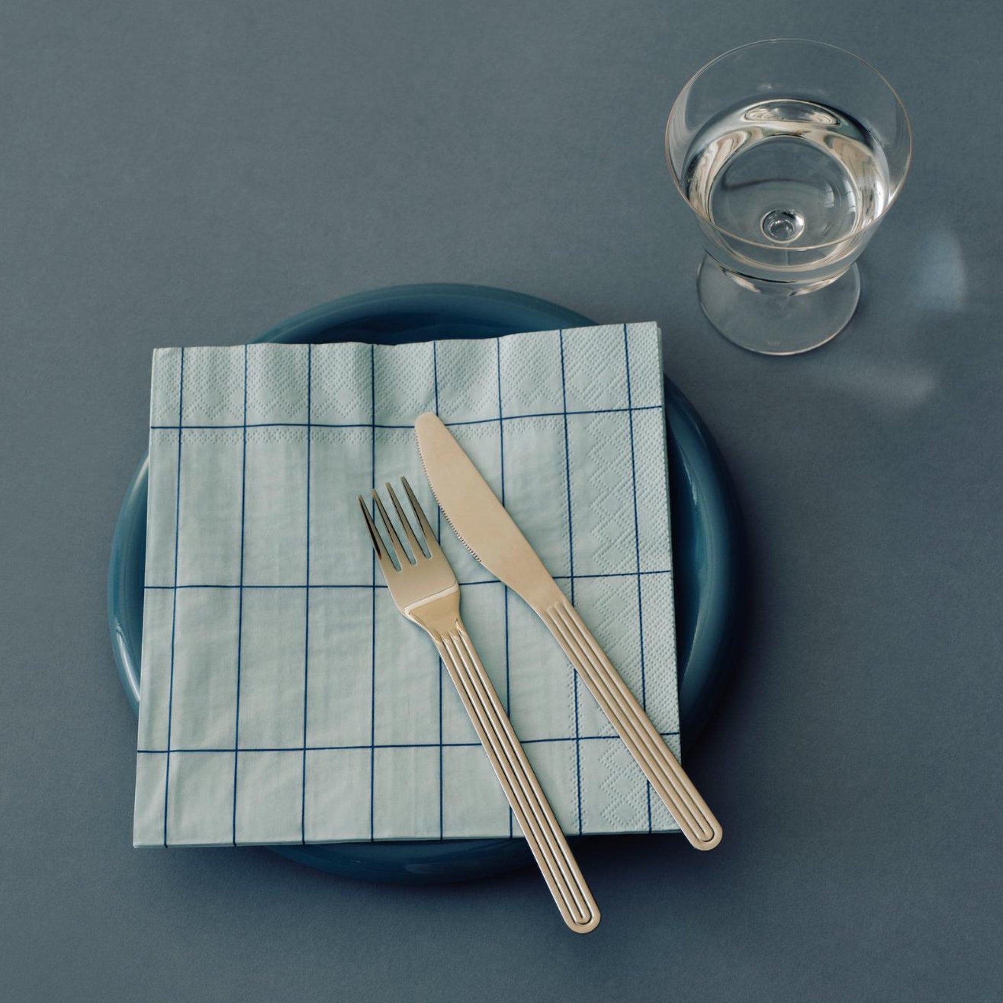 Een blauw bord met Hay Pattern Papieren Servetten Lichtblauw Blauw Grid 25/25 van Hay, met daarop vork en mes. Vlakbij staat een helder glas water op een grijsblauw oppervlak.