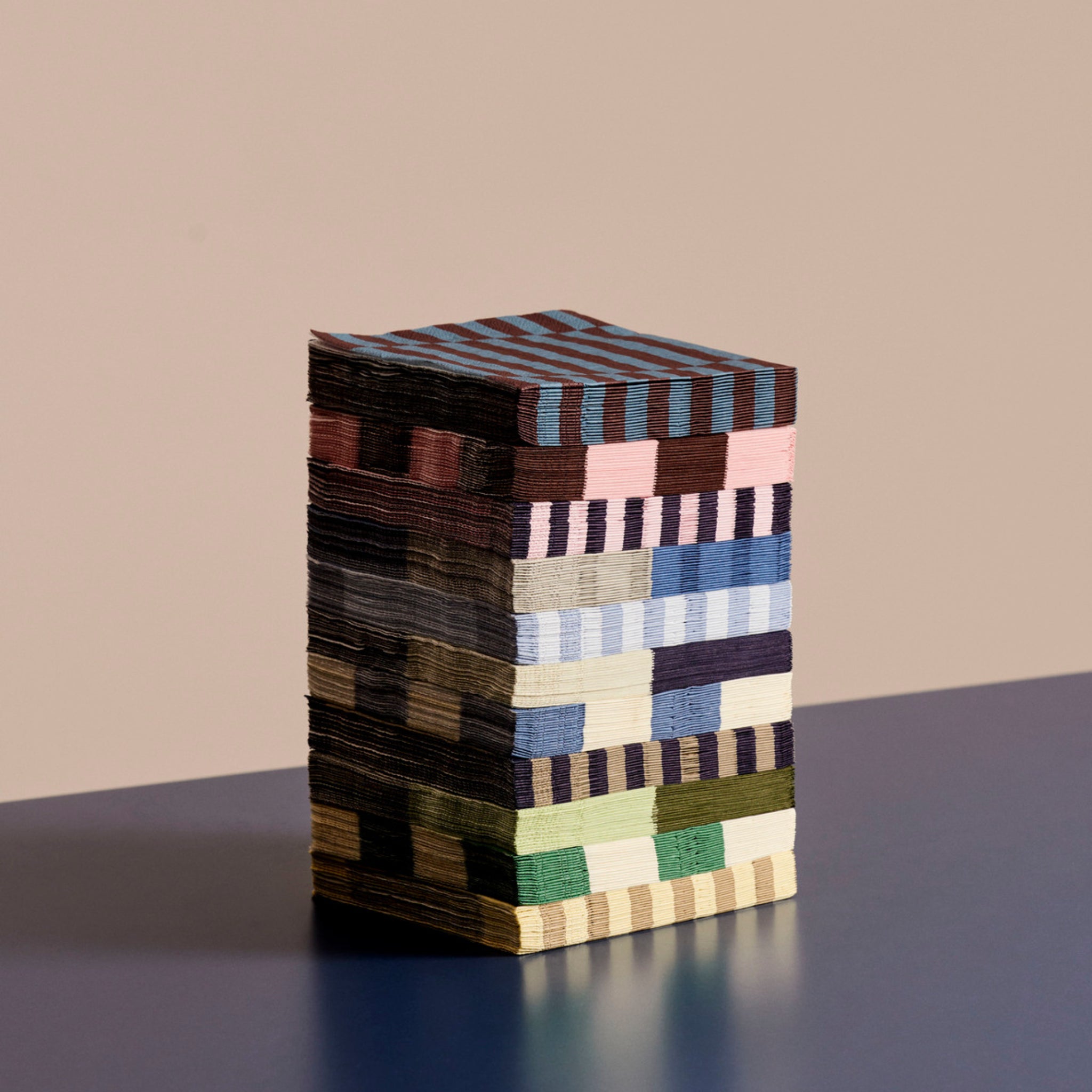 Een stapel Hay Pattern Papieren Servetten Grijs Blauw Stripe 33/33 servetten van Hay, met kleurrijke geometrische ontwerpen, is gerangschikt op een blauw oppervlak met een beige achtergrond.