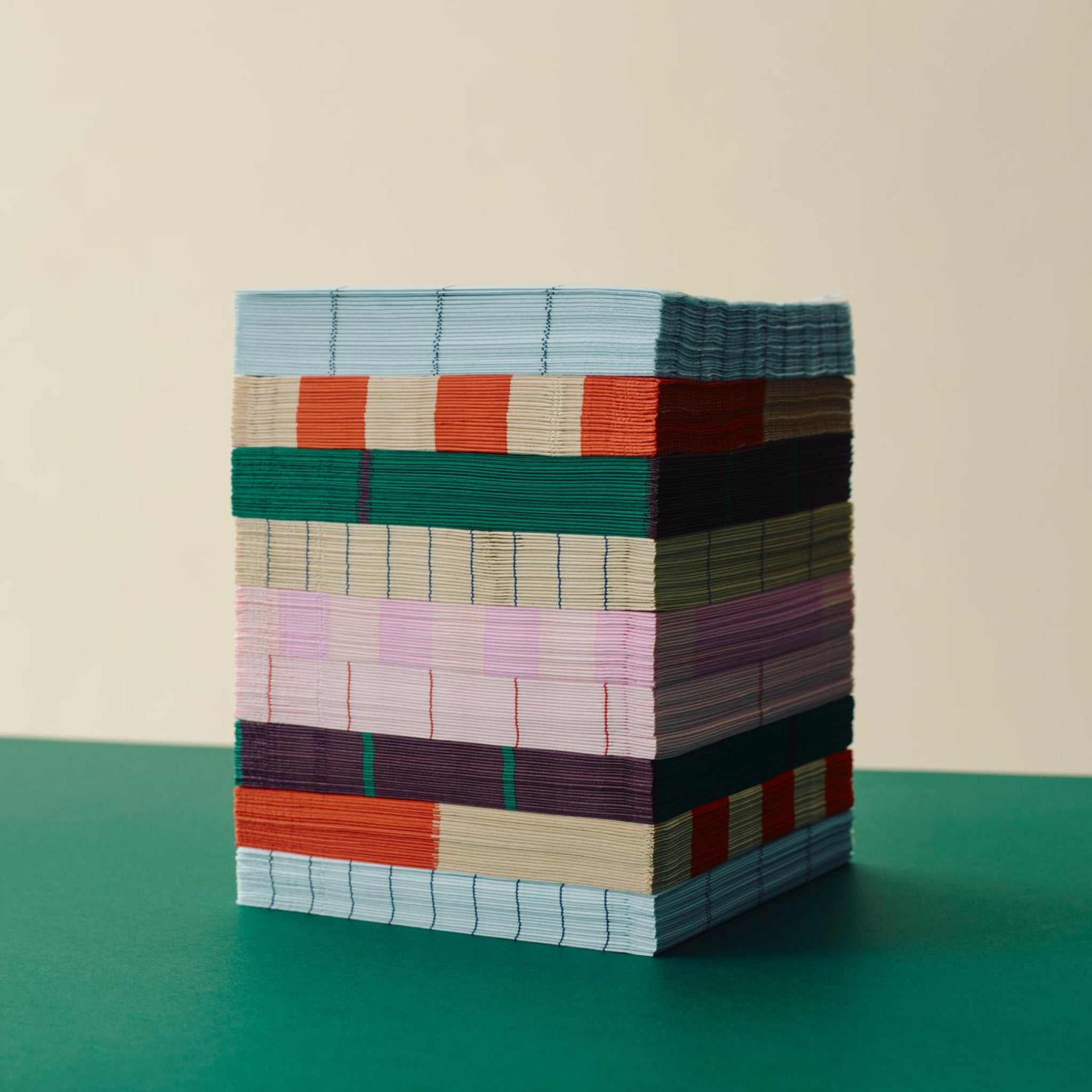 Een kubus van Hay Pattern Papieren Servetten Lichtroze Roos 33/33 van Hay, netjes gestapeld op een groen oppervlak tegen een beige achtergrond, laat levendige blauwe, rode, roze, groene en beige banden zien.