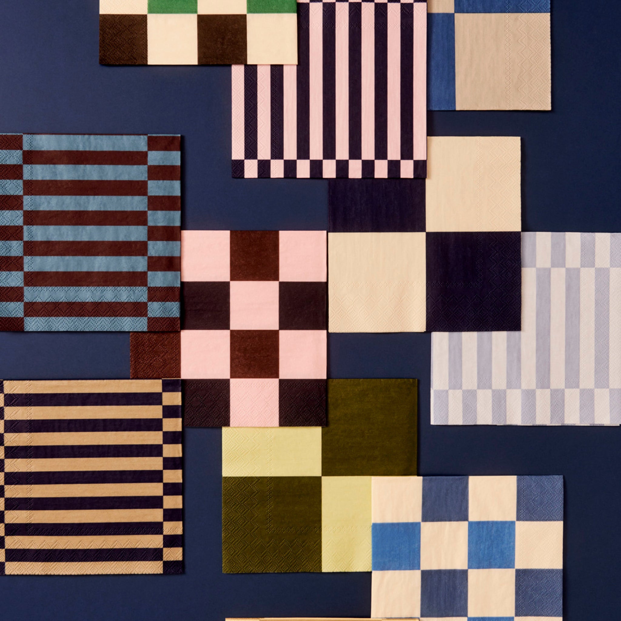 Patroon Papieren Servetten Off-white Blauw Ruit 33/33 van Hay is gerangschikt op een donkerblauwe achtergrond. De vierkante servetten hebben een blauw geruit patroon op een gebroken witte ondergrond.