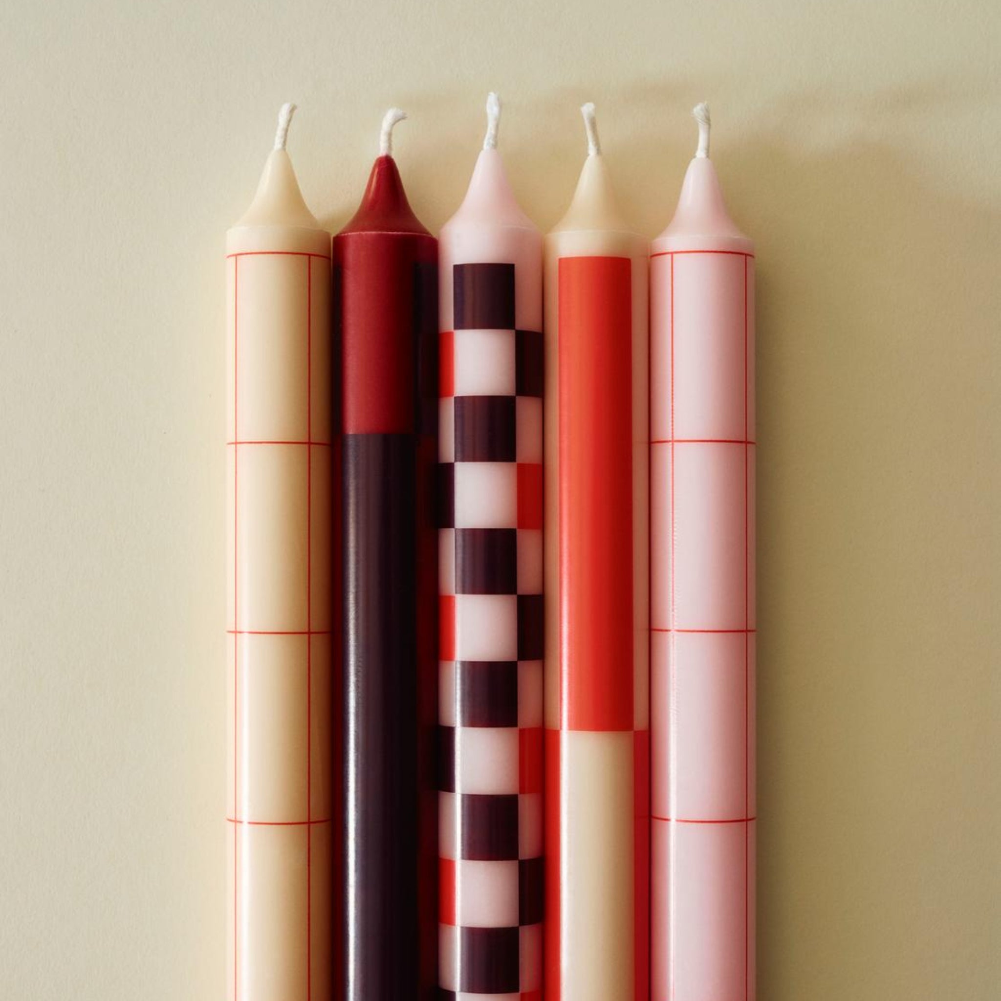 De Hay Patroon Kaarsen Rood en Bordeaux set bestaat uit vier elegante kaarsen met geometrische patronen in rood en bordeaux, naast elkaar gerangschikt op een lichtbeige achtergrond voor een stijlvolle touch.