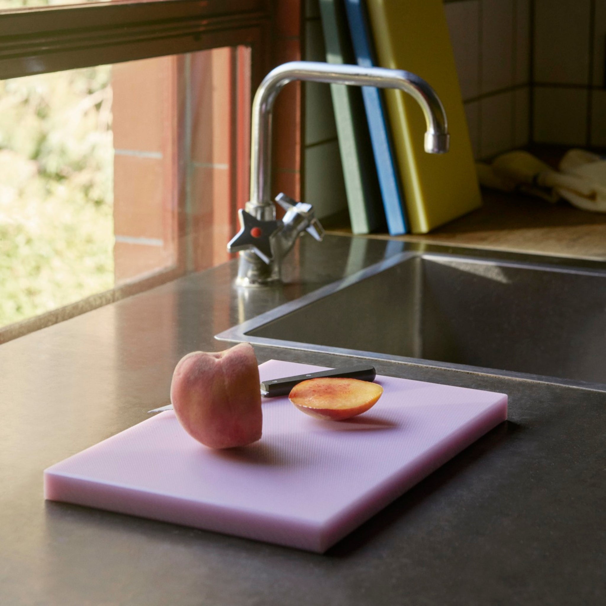 Een doormidden gesneden perzik en een mes rusten op een roze Hay Slice Snijplank Lavendel 20/30 bij een gootsteen in een zonovergoten keuken, terwijl drie rechtopstaande snijplanken, waaronder een van Hay, zichtbaar zijn op de achtergrond.