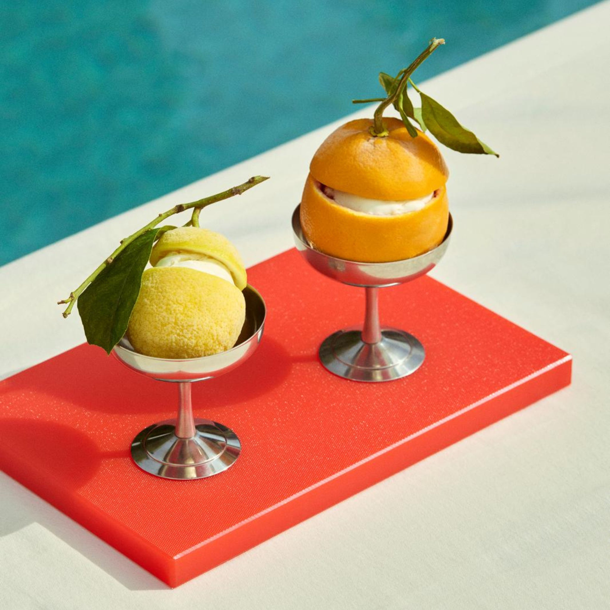 Twee metalen dessertbekers met geel en oranje ijs met citrusthema, elk bedekt met bladeren, rusten op een helderrode Hay Slice Snijplank Rood 20/30 van Hay, naast een sprankelend zwembad.