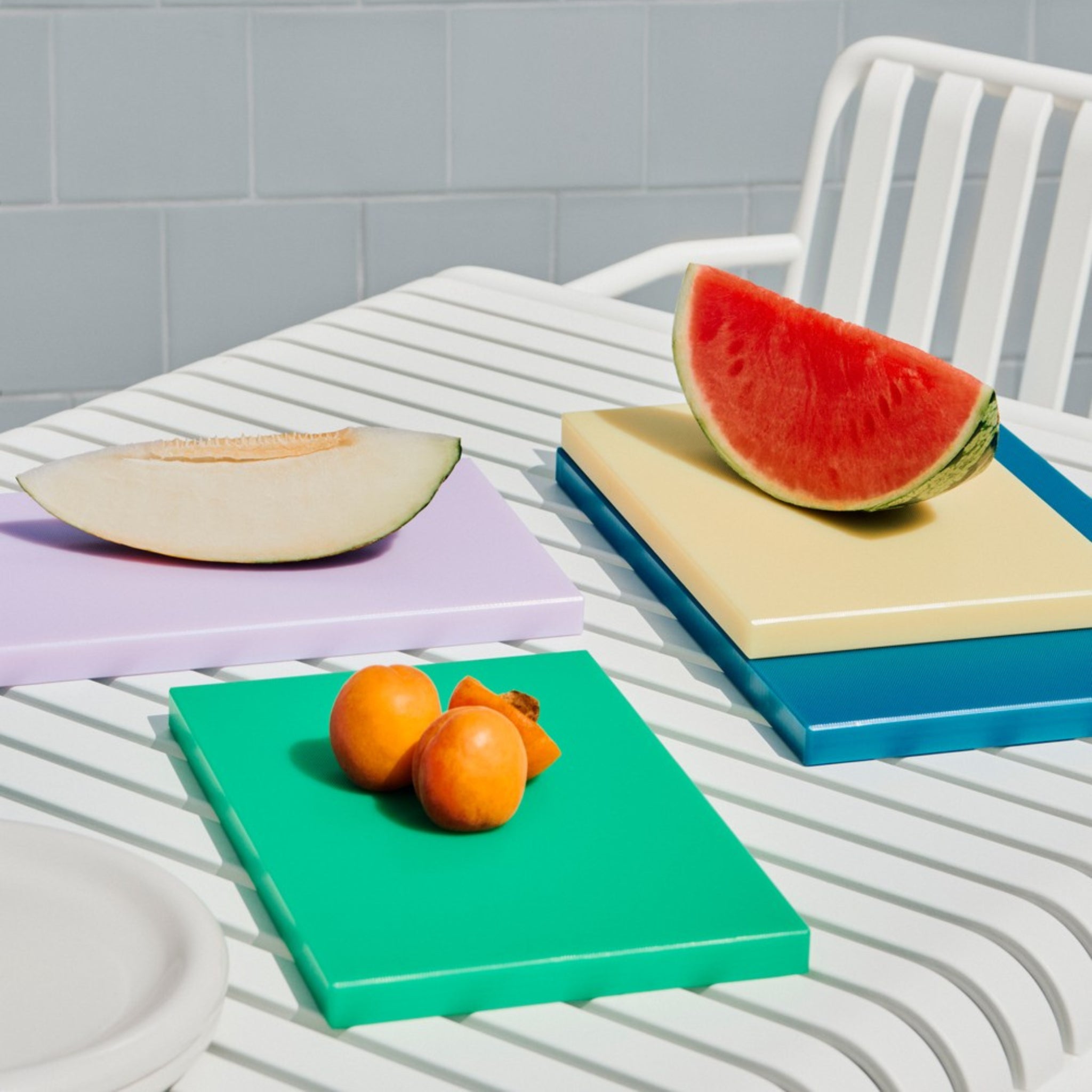 Een Hay Slice Snijplank Donkerblauw 25/38 snijplank van Hay, belegd met gesneden meloen en ander fruit, rust op een wit gestreepte buitentafel naast een witte stoel tegen een licht betegelde achtergrond.