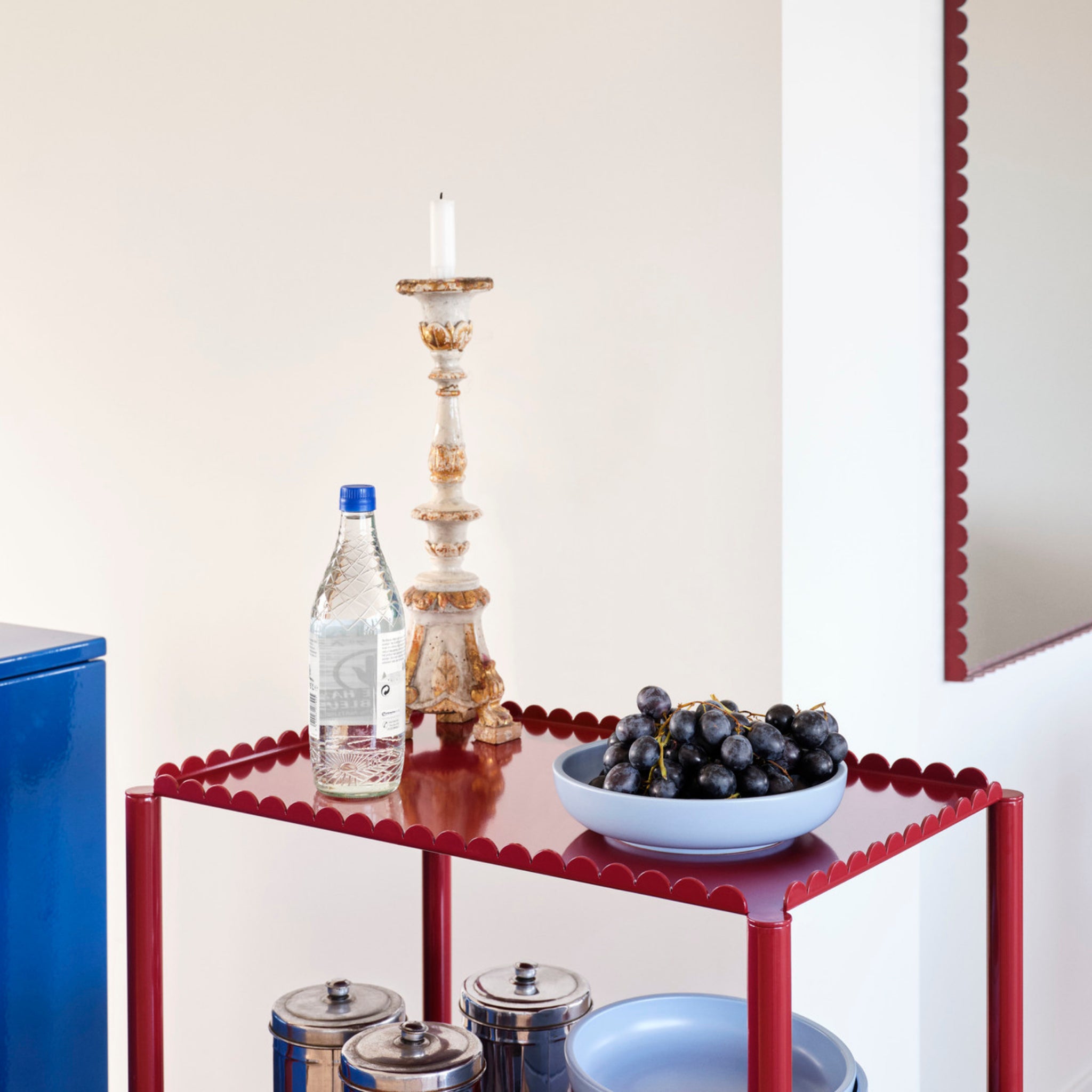 Een rood metalen karretje bevat een fles water, een sierlijke kandelaar met een witte kaars en de Hay Sobremesa Serveerschaal Lichtblauw D23 gevuld met zwarte druiven. Eronder staan blauwe schalen en metalen bussen. Aan de muur hangt een geschulpte spiegel.
