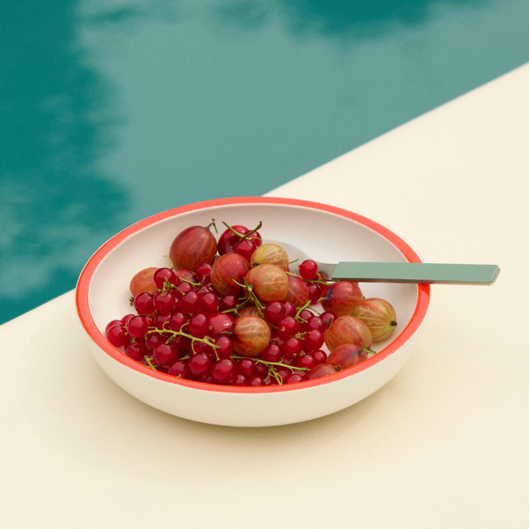 De Hay Sobremesa Serveerschaal Wit, Rode Rand D20 van Hay, gevuld met rode kruisbessen en aalbessen, ligt op een bleek oppervlak bij een turquoise poel; een lepel rust in deze elegante witte kom met een rode rand.