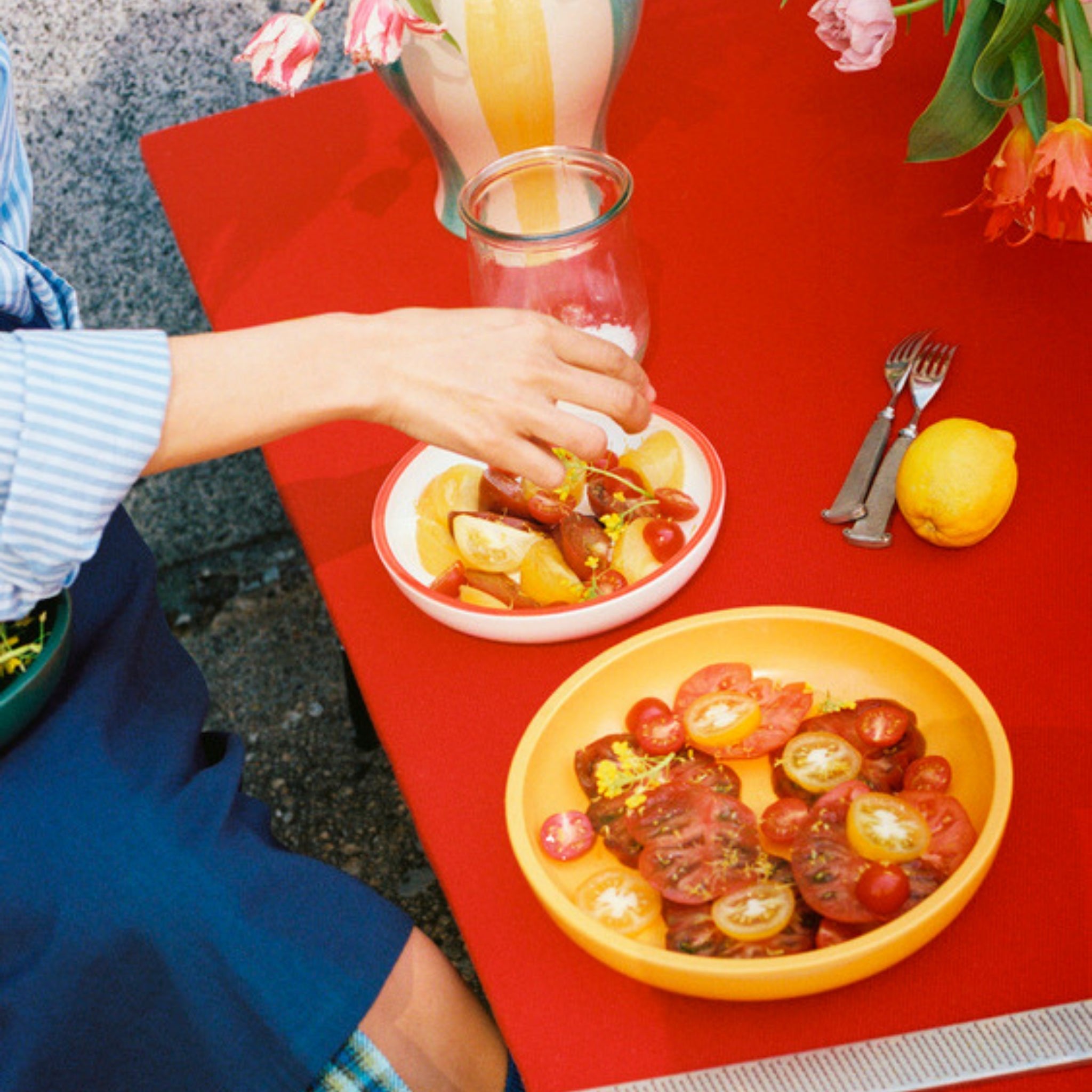 Een persoon aan een rode tafel reikt naar een schaal met gesneden tomaten en citroenen op de Hay Sobremesa Serveerschaal Geel D25 van Hay, met een ander kleurrijk tomatensaladebord ervoor, bestek en een hele citroen naast een vaas met bloemen.
