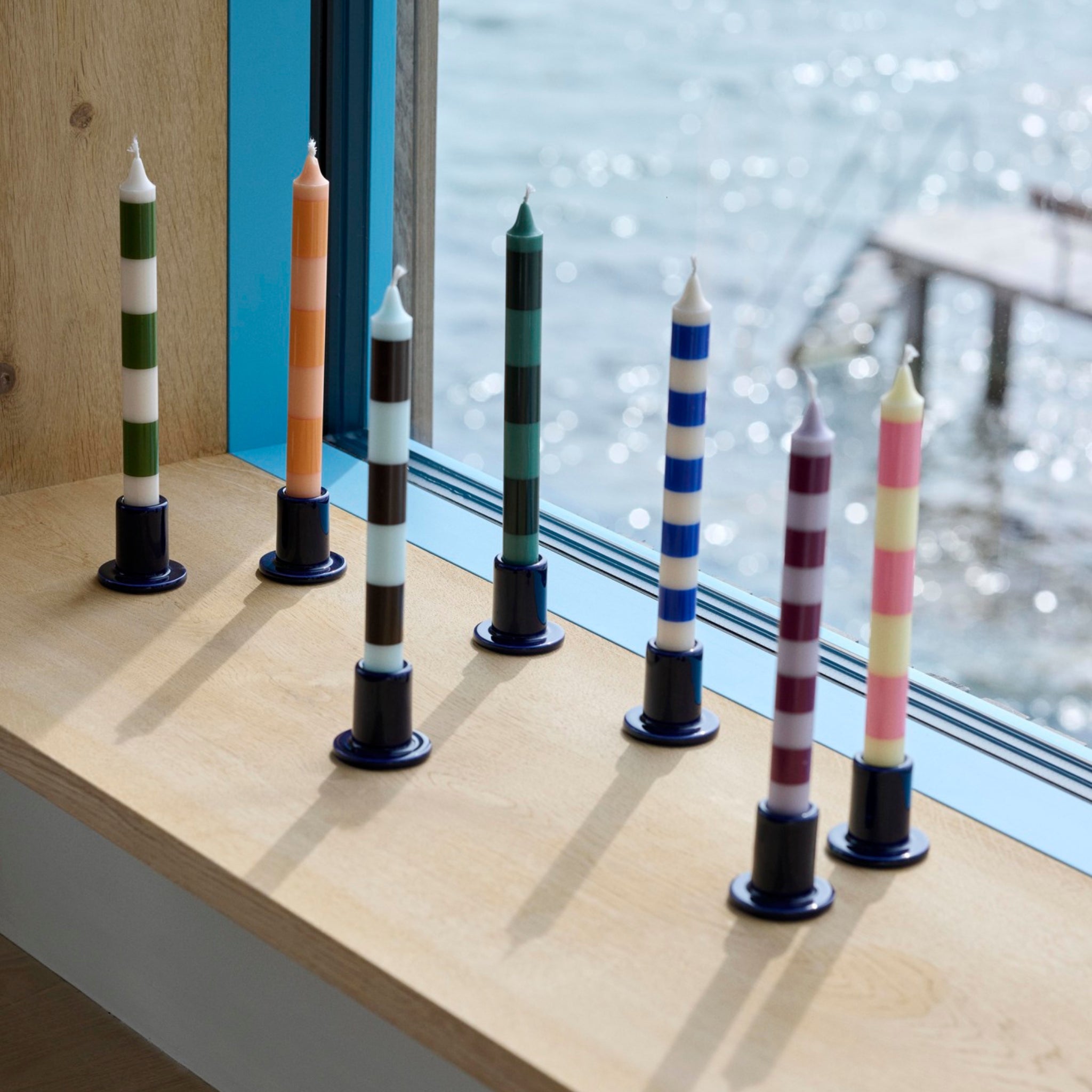 Zes gestreepte kaarsen in Hay Tube Kandelaar Middernachtblauw houders van Hay staan op een houten vensterbank, zonlicht werpt schaduwen en water zichtbaar buiten, met het moderne blauwe aardewerk voor een stijlvolle touch.