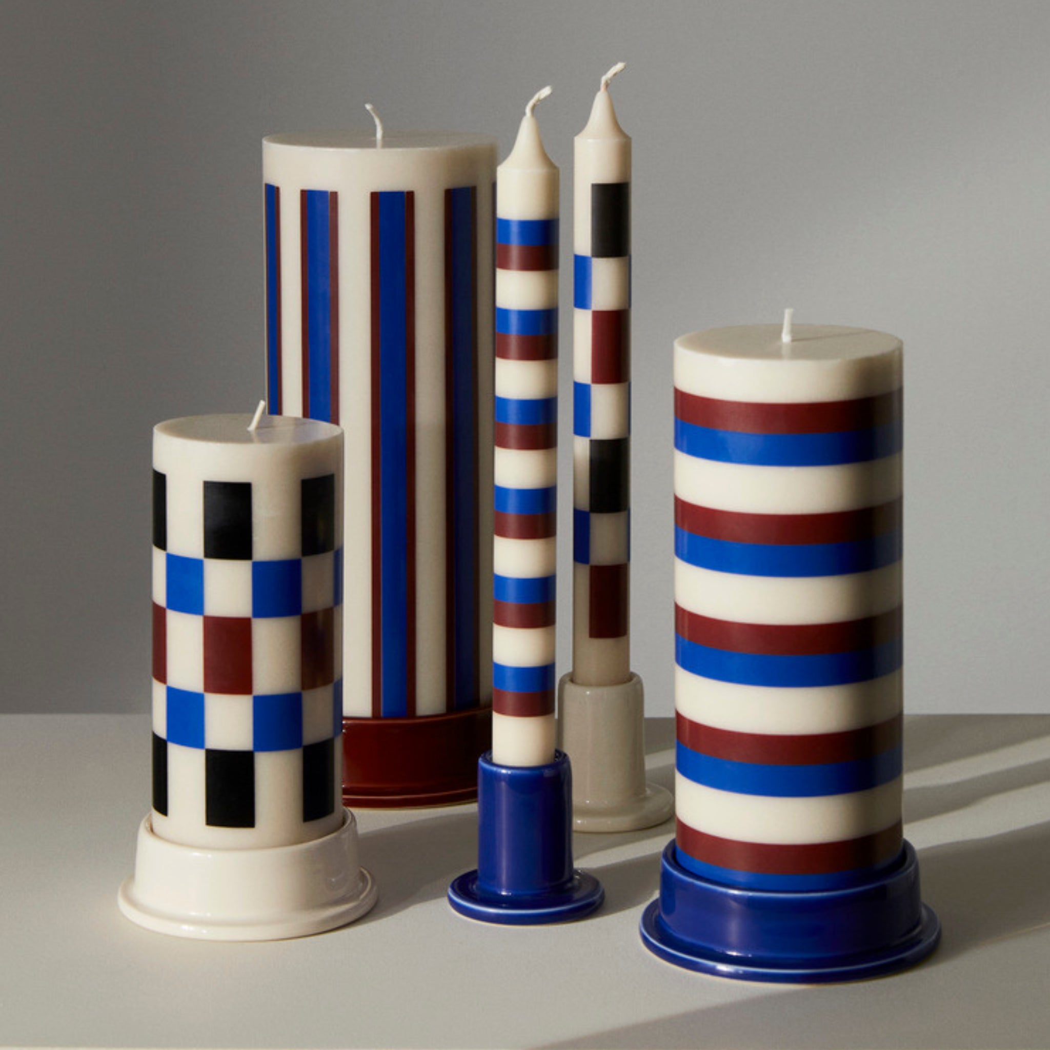 Hay Tube Kandelaar Middernachtblauw van Hay heeft vier geometrische pilaar- en conuskaarsen in blauwe, rode en zwarte aardewerk houders, rechtopstaand op een neutraal oppervlak tegen een zacht verlichte grijze achtergrond voor een moderne touch.