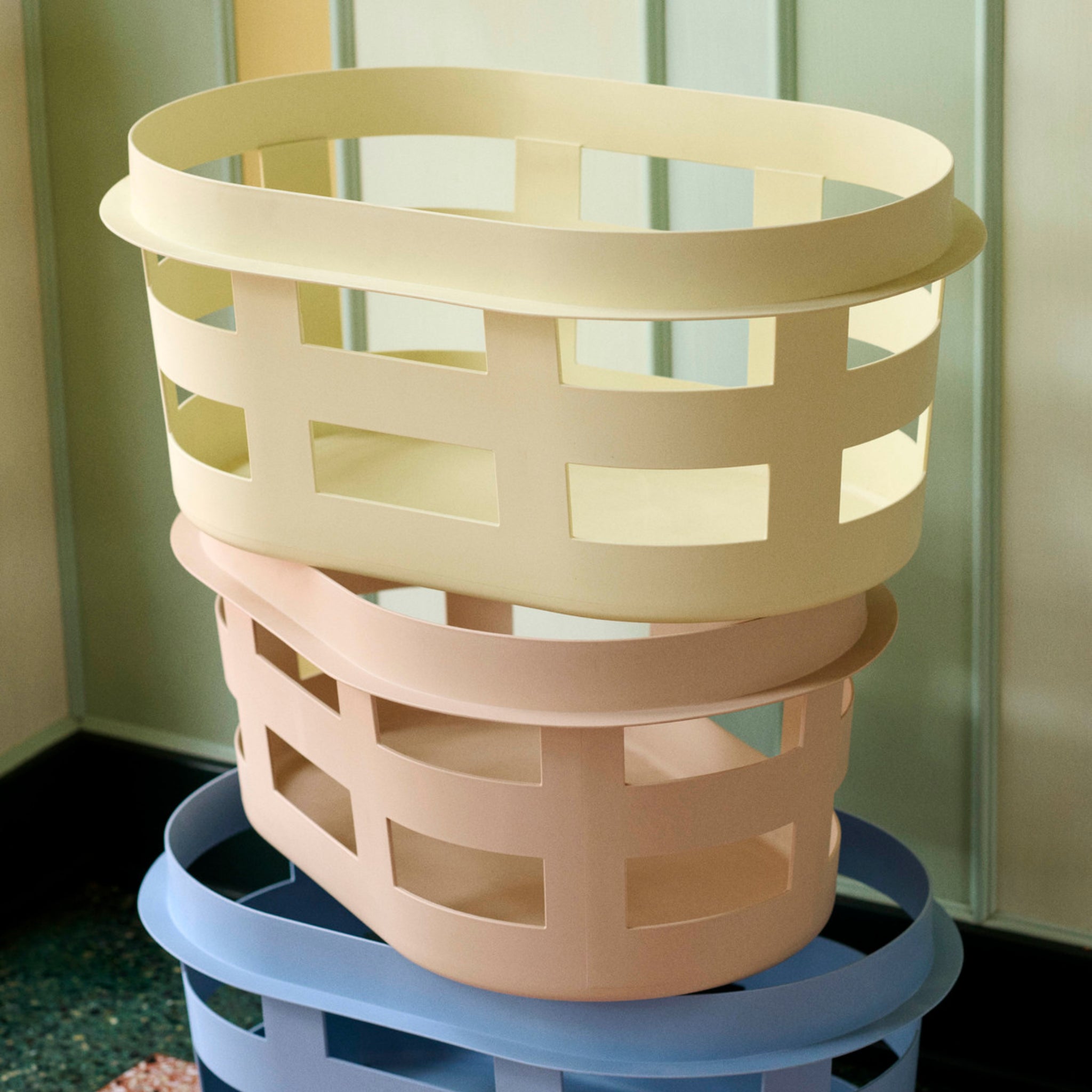 Drie moderne, rechthoekige Hay Wasmand Geel wasmanden van Hay - pastelgeel, roze en blauw - staan netjes gestapeld tegen een lichtgroene muur en een donkerkleurige vloer.