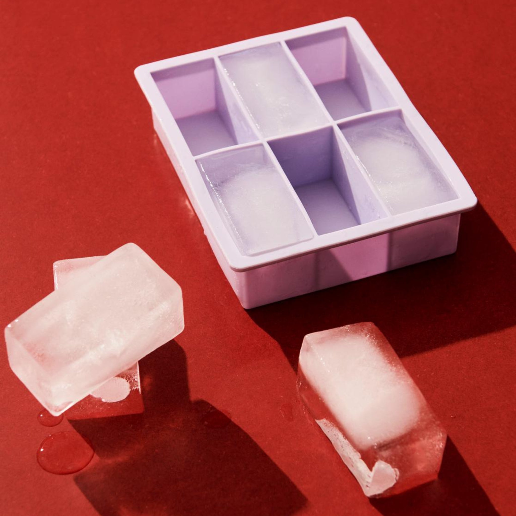 De Hay ijsblokjesvorm Rechthoekig XXL Lila staat op een rood oppervlak, met vijf grote rechthoekige compartimenten, met twee heldere ijsblokjes ernaast en een klein plasje gesmolten water vlakbij.