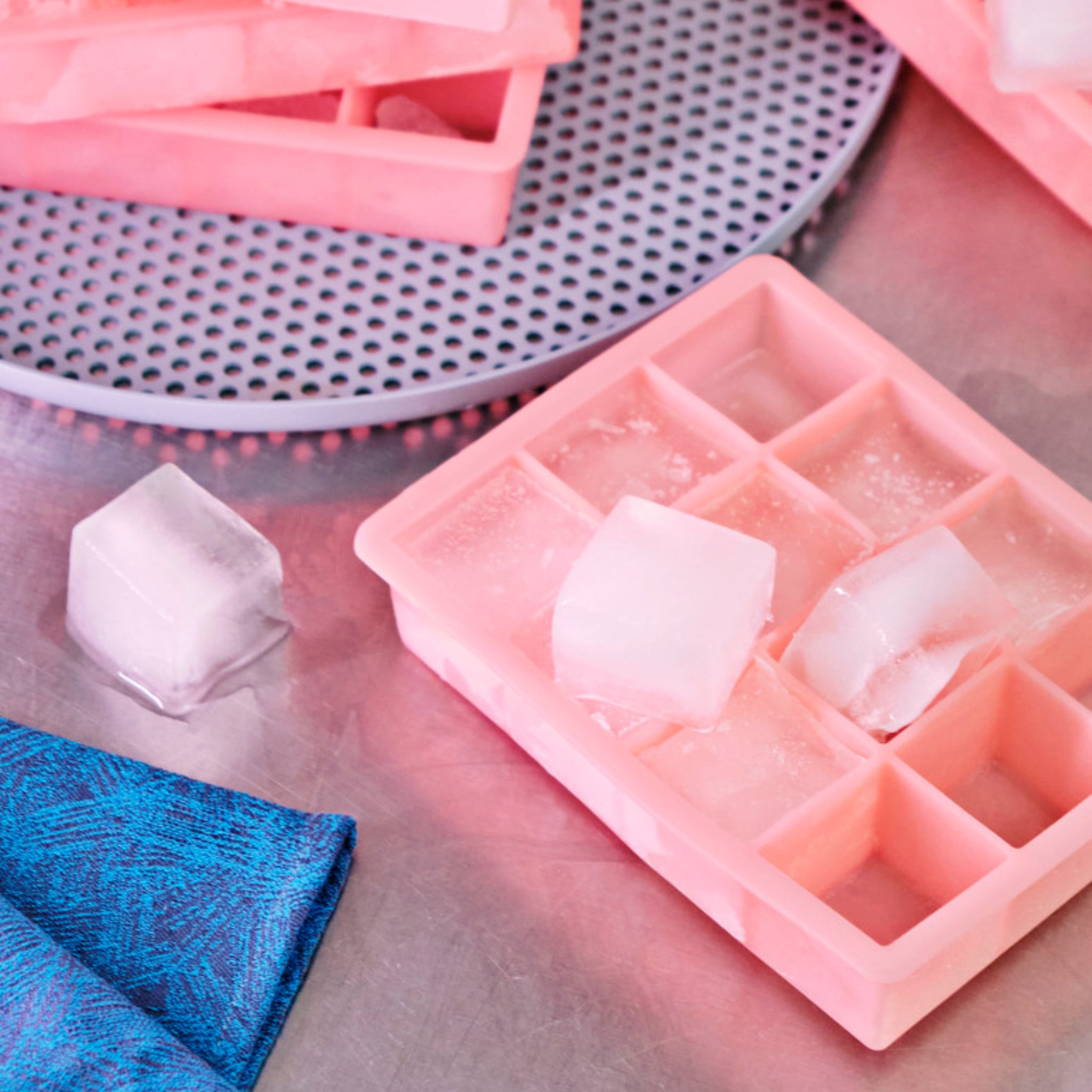 Een Hay ijsblokjesvorm Vierkant XL Lichtblauw zit op een metalen oppervlak bij een blauw gedessineerd doek, met extra lichtblauwe bakjes en een geperforeerd rond dienblad op de achtergrond.