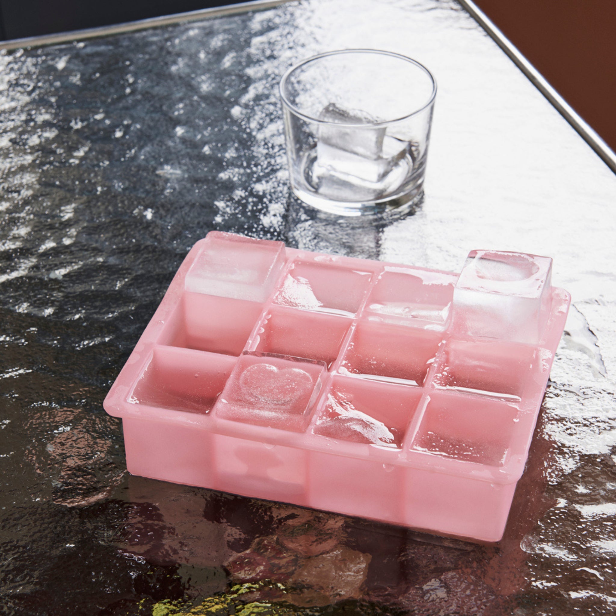De Hay ijsblokjesvorm Vierkant XL Roze van Hay, een roze siliconen ijsblokjesbak voor XL blokjes, staat op een reflecterende glazen tafel met een glas grote ijsblokjes erachter.