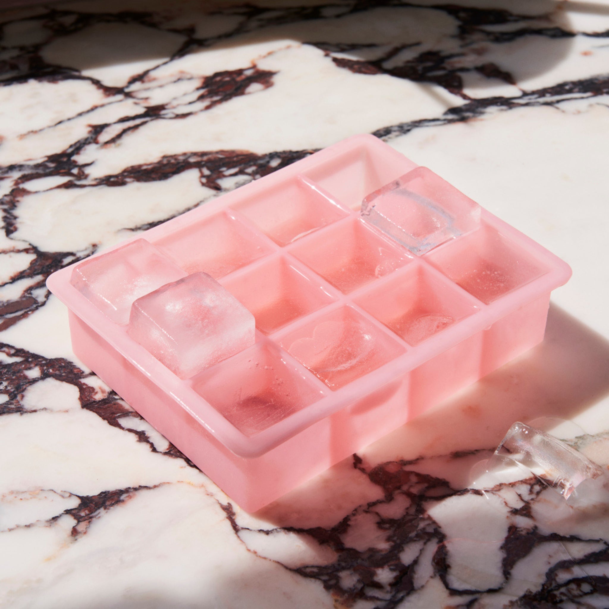 De Hay ijsblokjesvorm Vierkant XL Roze van Hay rust op een wit en donker gemarmerd oppervlak in fel zonlicht, met verschillende XL ijsblokjes verspreid naast het roze siliconen dienblad.