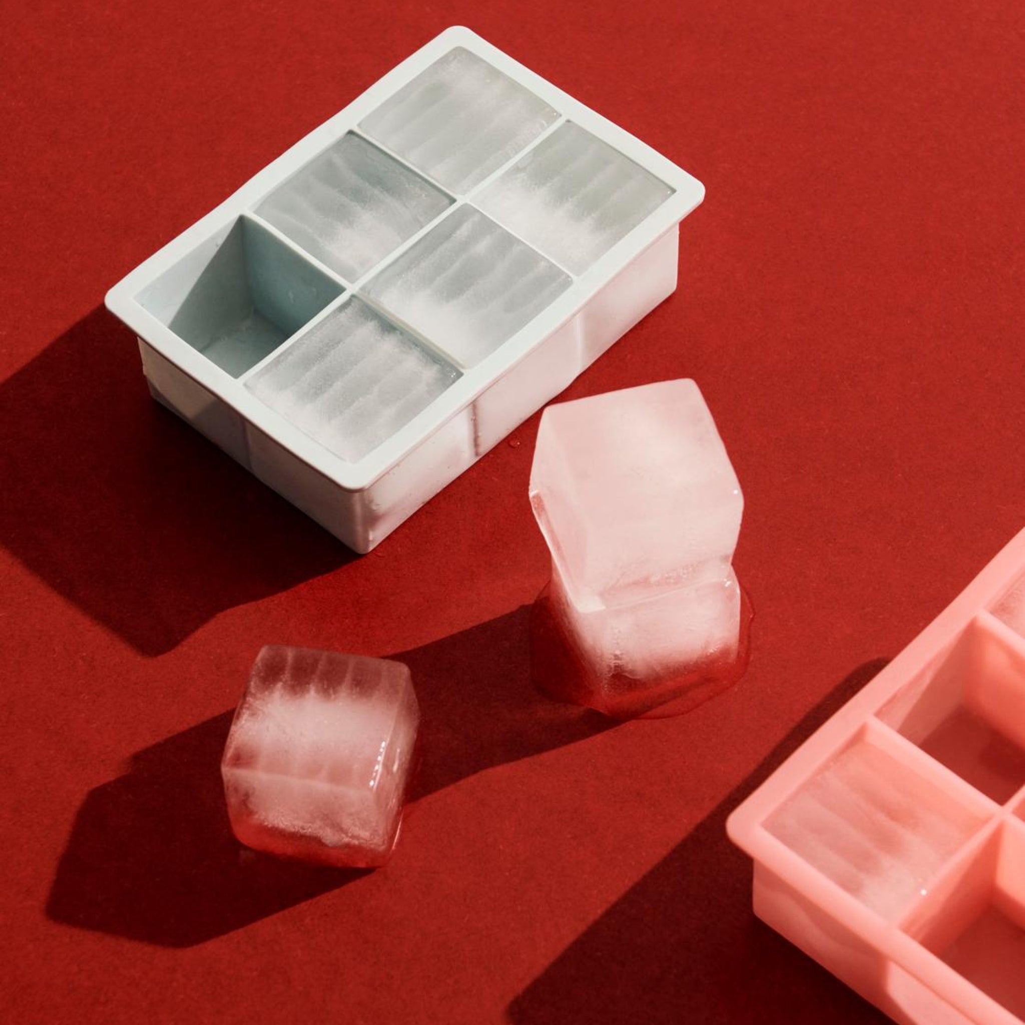 Een Hay ijsblokjesvorm Vierkant XXL Ijsblauw van Hay en twee XXL ijsblokjes rusten op een rood vlak, met een roze dienblad gedeeltelijk zichtbaar in de hoek. Heldere verlichting creëert duidelijke schaduwen.