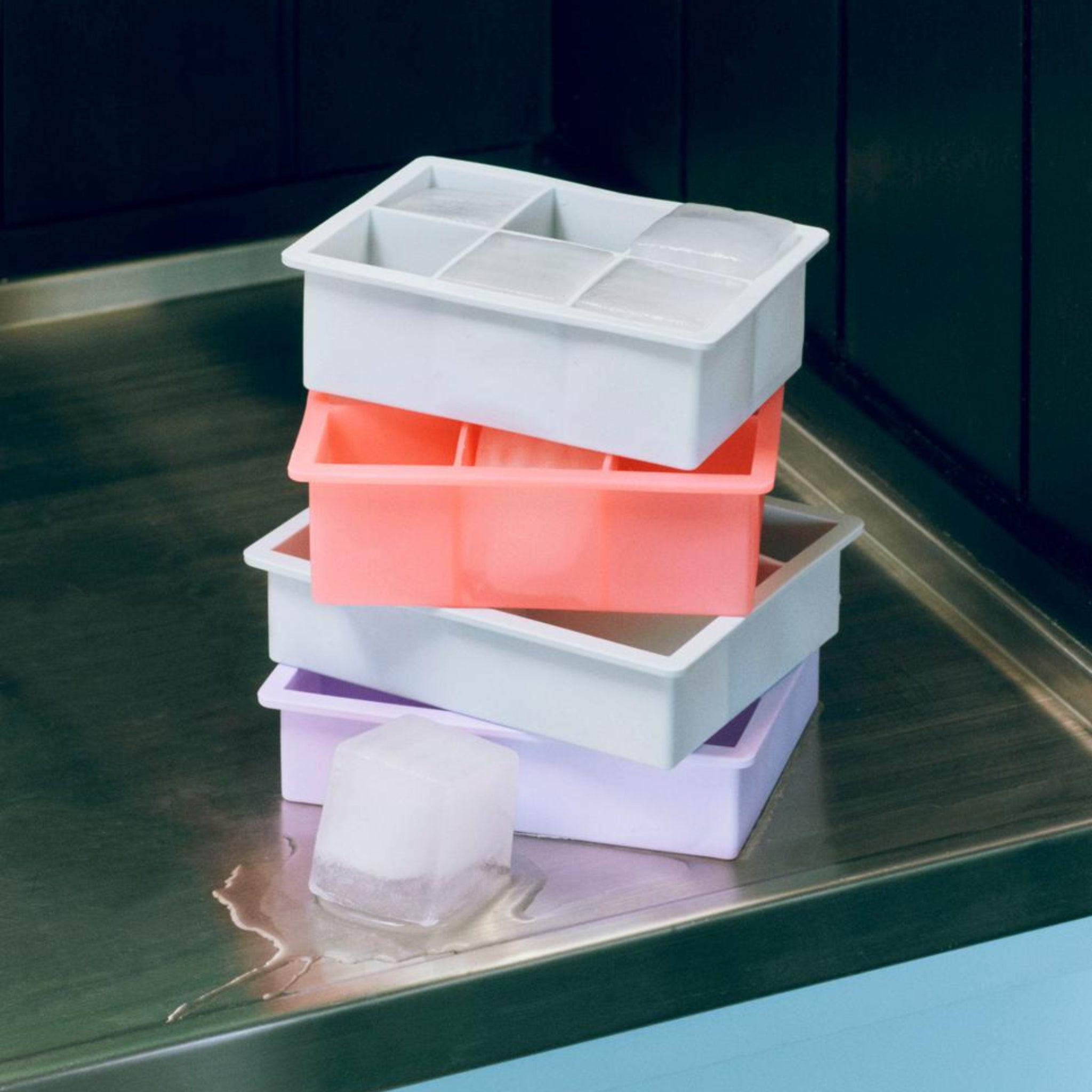 Een Hay ijsblokjesvorm Vierkant XXL in ijsblauw staat gestapeld met andere bakjes op een roestvrijstalen oppervlak, sommige met XXL ijsblokjes. Eén groot vierkante ijsblokje van het Hay dienblad is gevallen en ligt te smelten op het aanrecht.