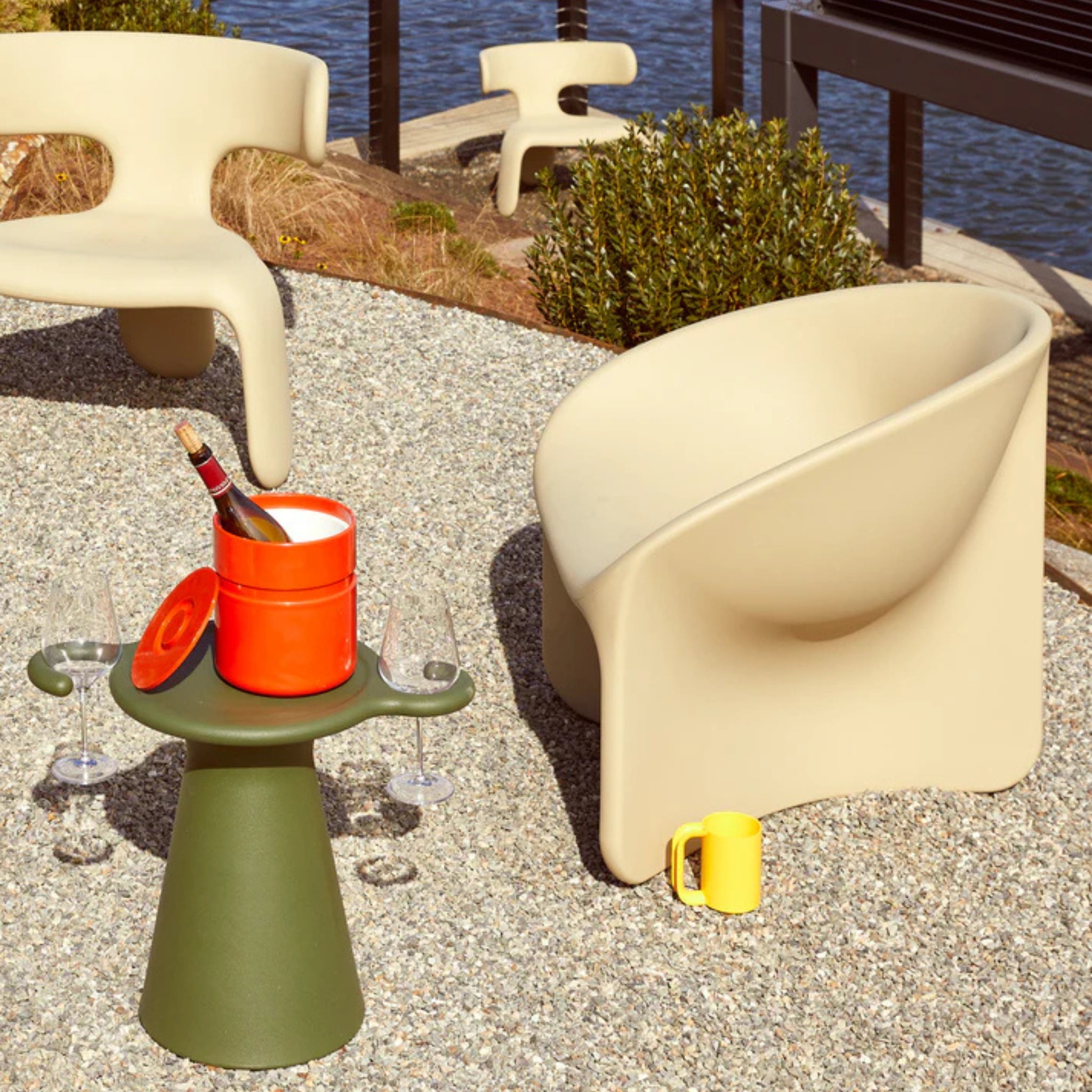 Een moderne patio met gemodelleerde beige stoelen en een groene bijzettafel met een Heller Asti Ijsemmer Oranje met wijn, twee glazen en een gele mok staat op grind in de buurt van water en groen.