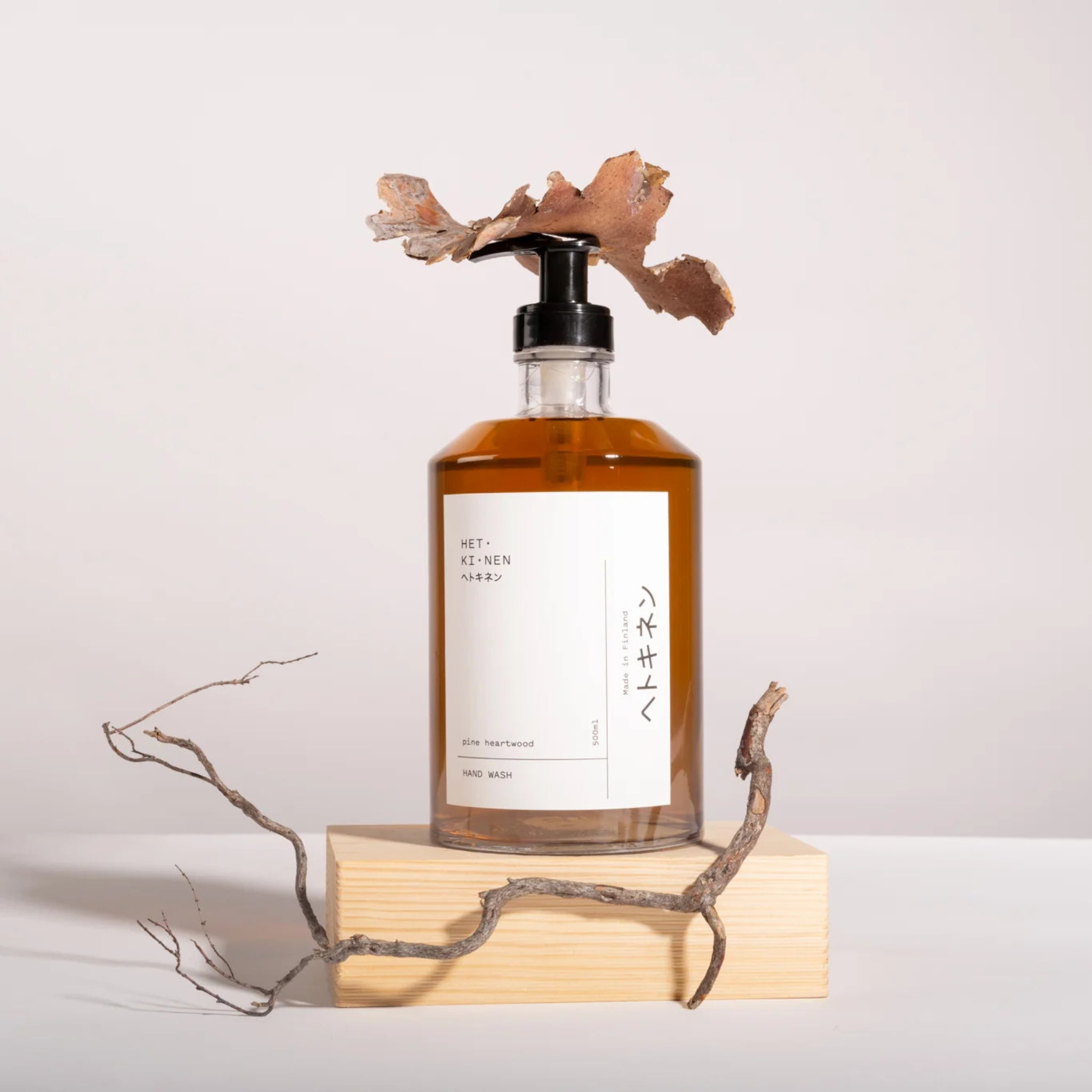 Een heldere pompfles Hetkinen Handzeep Pine Heartwood 500 ml met amberkleurige vloeistof staat op een houten blok met daarop een gedroogd blad, naast een kale tak. Minimalistische Japanse en Engelse labels accentueren deze natuurlijke zeep van Hetkinen.