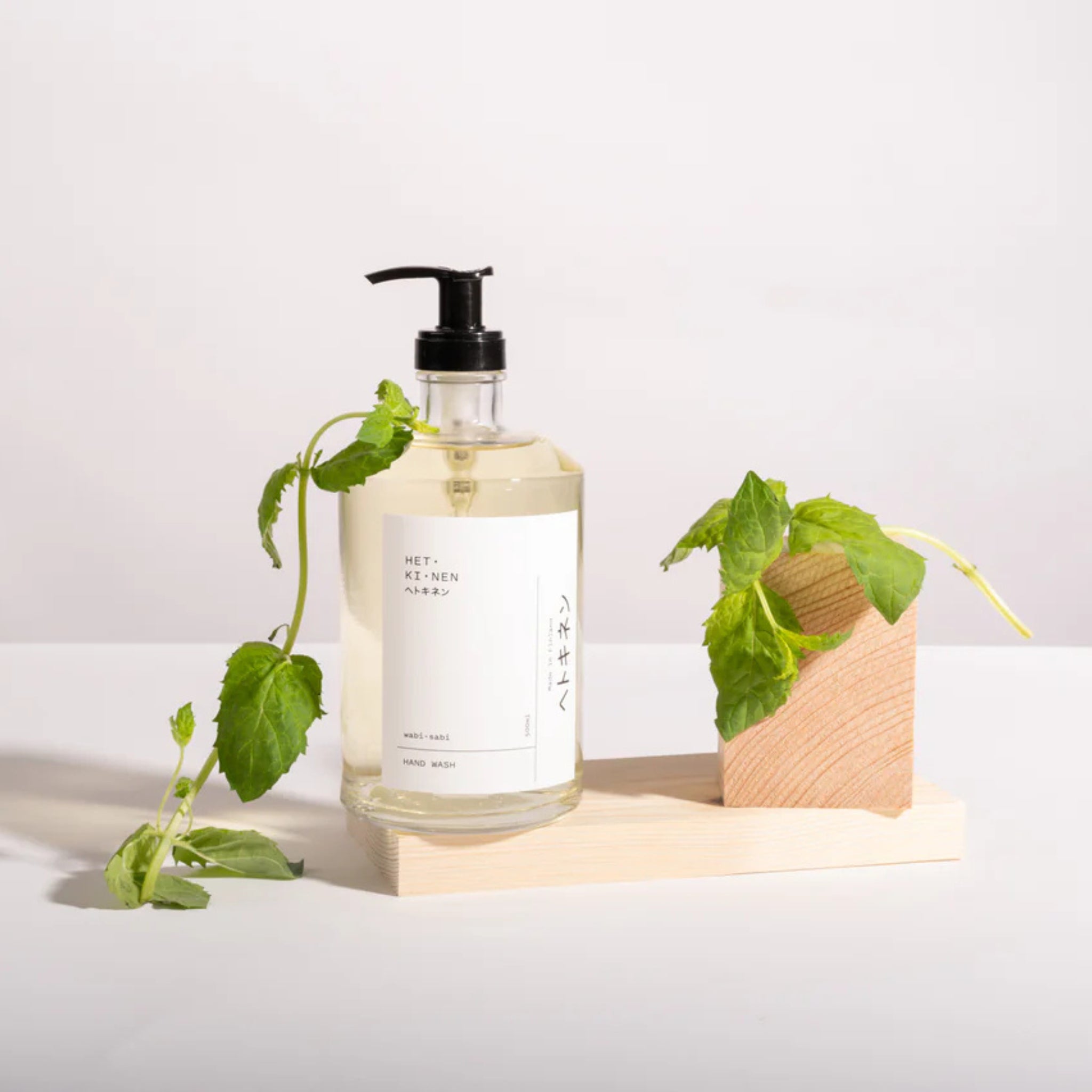 Een 500 ml heldere pompfles Hetkinen Handzeep Wabi-Sabi met een wit etiket staat op een licht houten dienblad naast verse groene muntblaadjes en een klein houten blokje, alles tegen een effen witte achtergrond.
