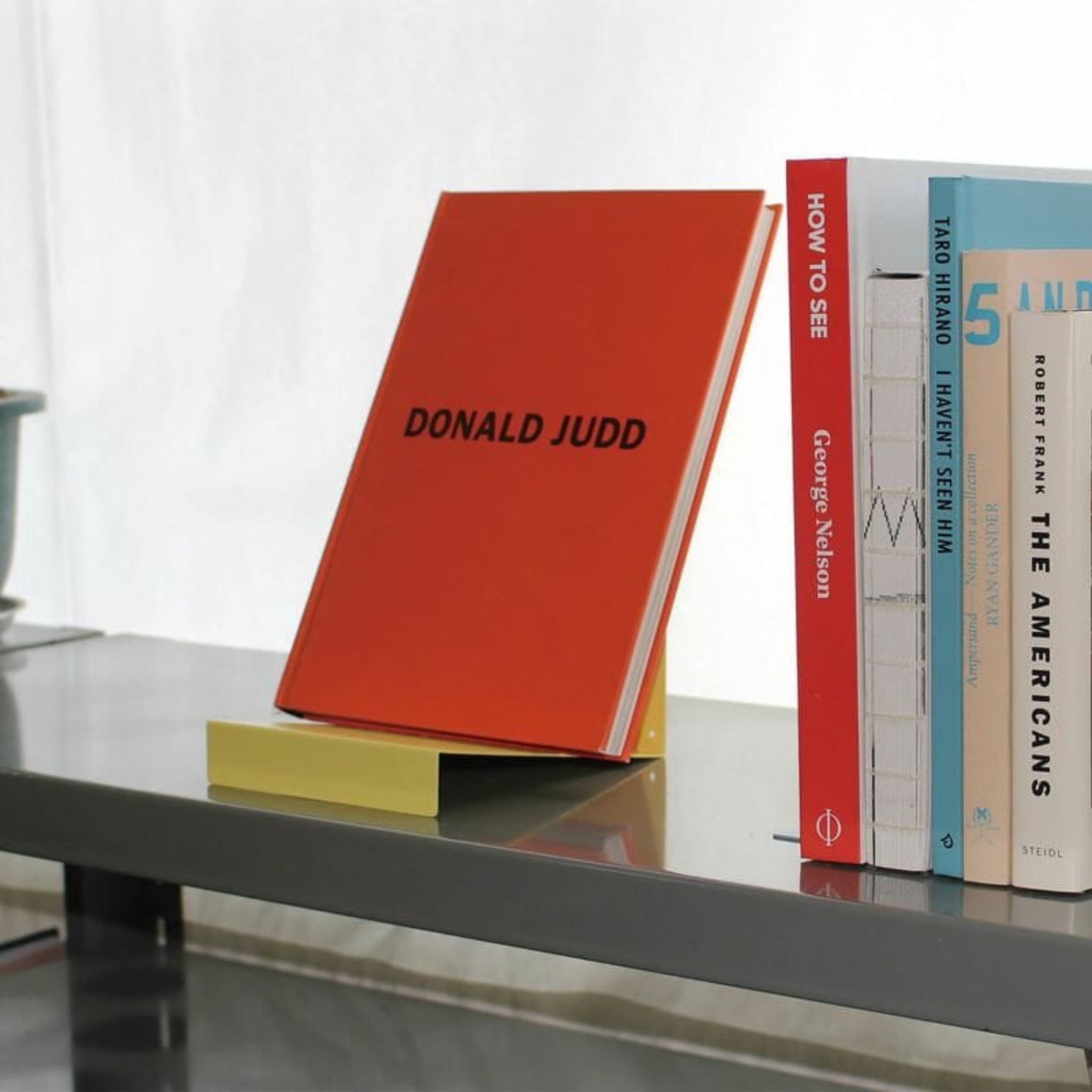 Een rood boek met de titel DONALD JUDD staat rechtop op een gele Hightide Boekenstandaard & Display Geel van Hightide, op een grijze plank naast boeken als HOW TO SEE en THE AMERICANS.