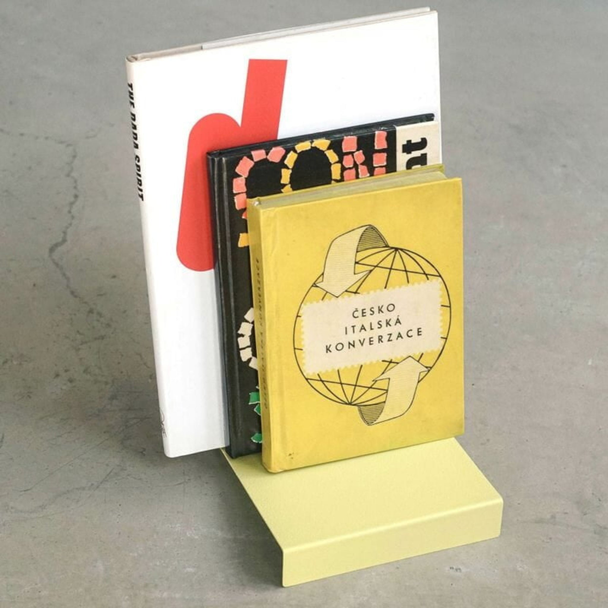 Vier boeken staan rechtop op een Hightide Boekenstandaard & Display Geel van Hightide. Het voorste boek heeft een gele kaft met een wereldbol en Tsjechische tekst; drie andere zijn gedeeltelijk zichtbaar erachter, allemaal tegen een grijze achtergrond.