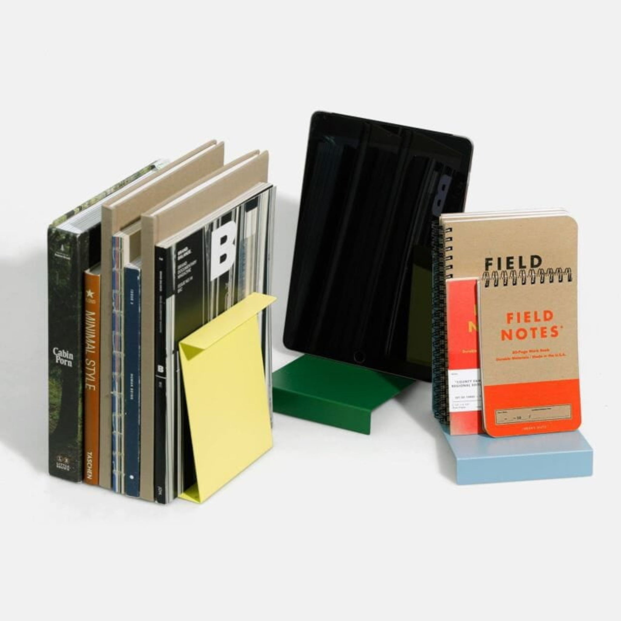 Een set boeken, notitieblokken - sommige met het label Field Notes - en een tablet staan netjes rechtop tussen de boekensteunen van Hightide Boekenstandaard & Display Geel, allemaal tegen een effen witte achtergrond.