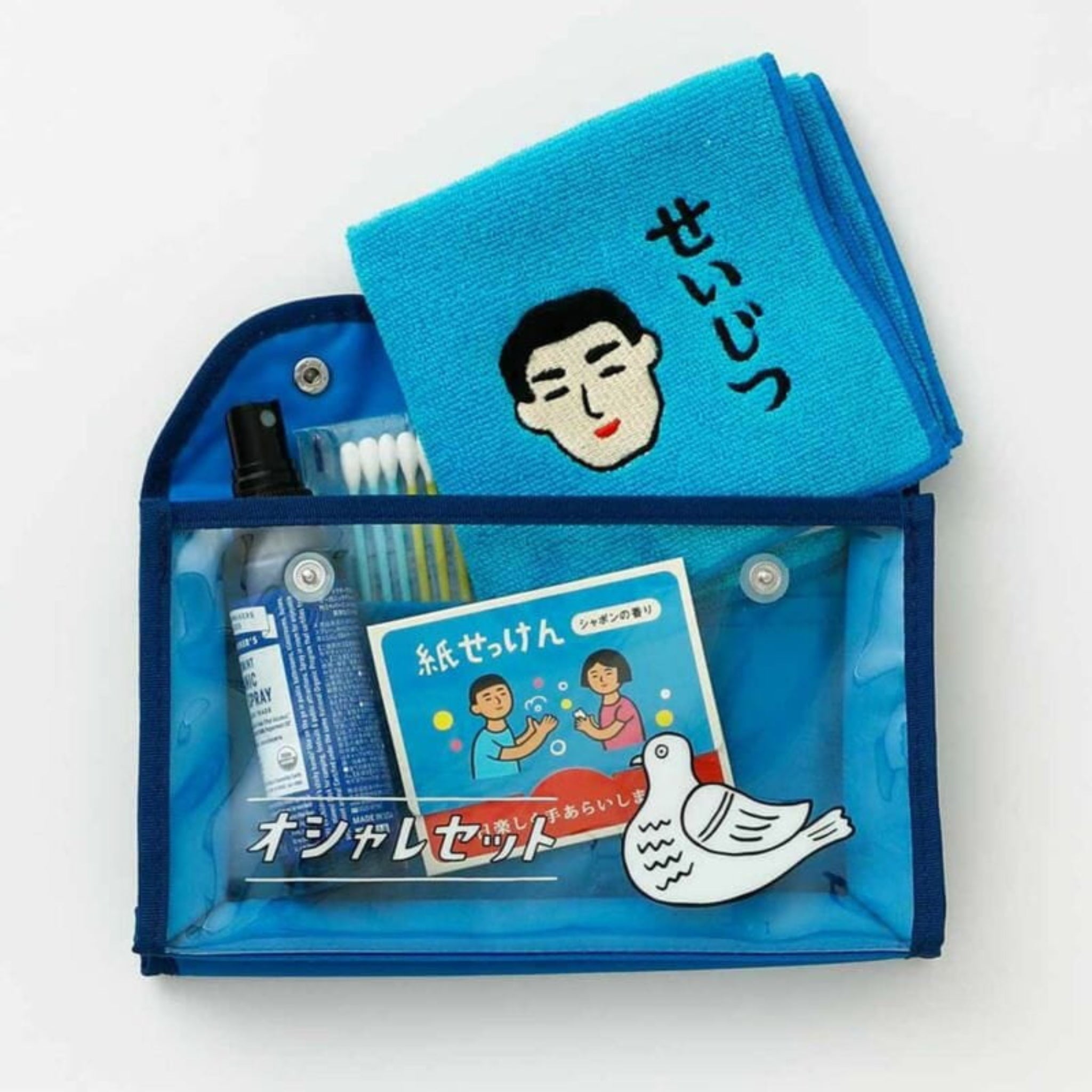 Een transparant blauw etui van Hightide bevat gevouwen Hightide New Retro Handdoek Blauw 25/25 met een cartoongezichtje en Japanse tekst, plus wattenstaafjes, een sprayflacon, een ansichtkaart met twee mensen en een duif, en een tube crème.