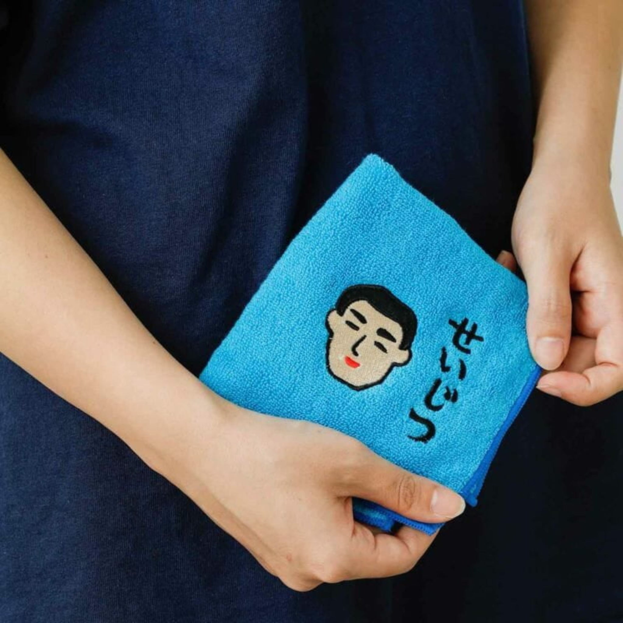 Een persoon in een donkerblauw shirt houdt de Hightide New Retro Handdoek Blauw 25/25 vast, een blauwe handdoek van het Japanse merk Hightide met een geborduurd gezicht en Japanse tekens.