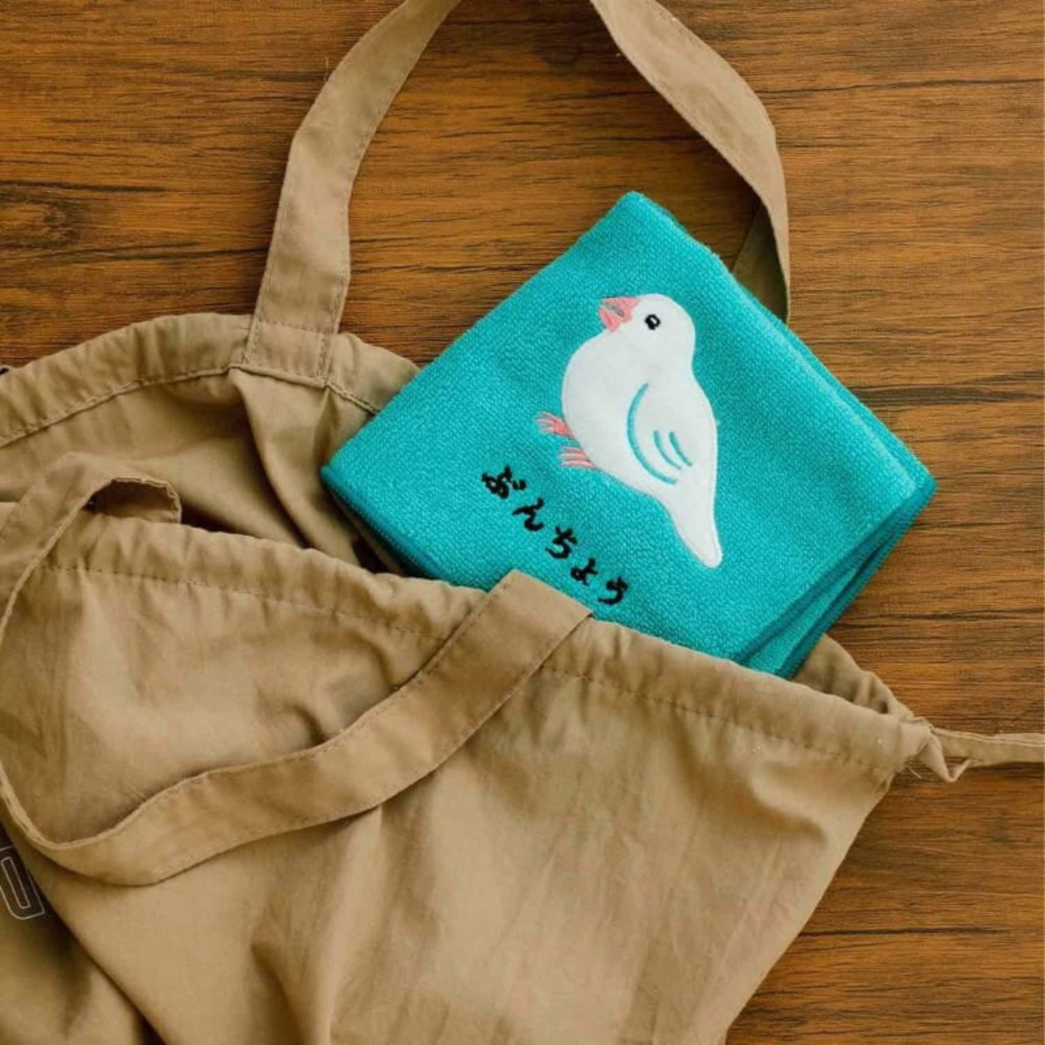 Een Hightide New Retro Handdoek Groen 25/25 van het Japanse merk Hightide zit in een bruine draagtas op een houten oppervlak, met een illustratie van een witte vogel met blauwe vleugels en Japanse tekst eronder.