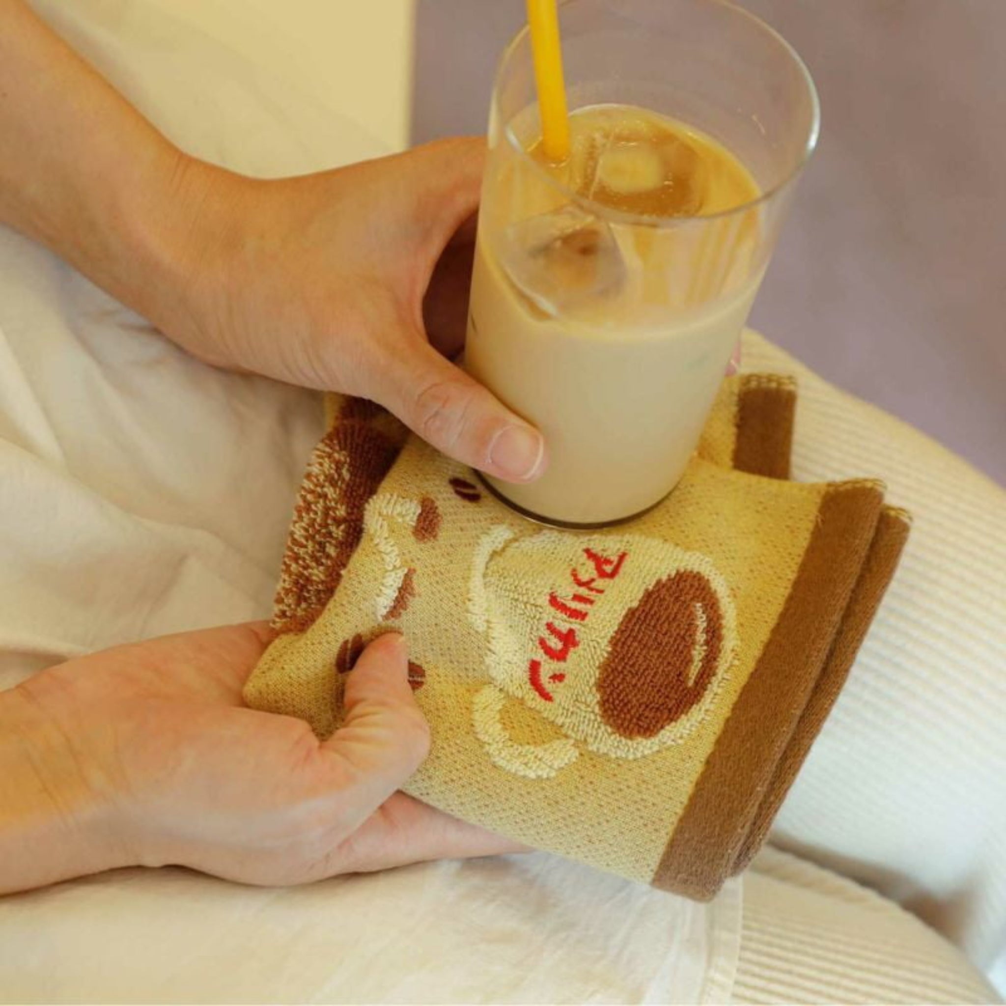 Een persoon houdt een glas ijskoffie met een geel rietje in de ene hand en een beige Hightide Handdoek Koffie 25/25 van Hightide, met een schattig berengezicht en het woord "ruffle" in rood, in de andere hand.