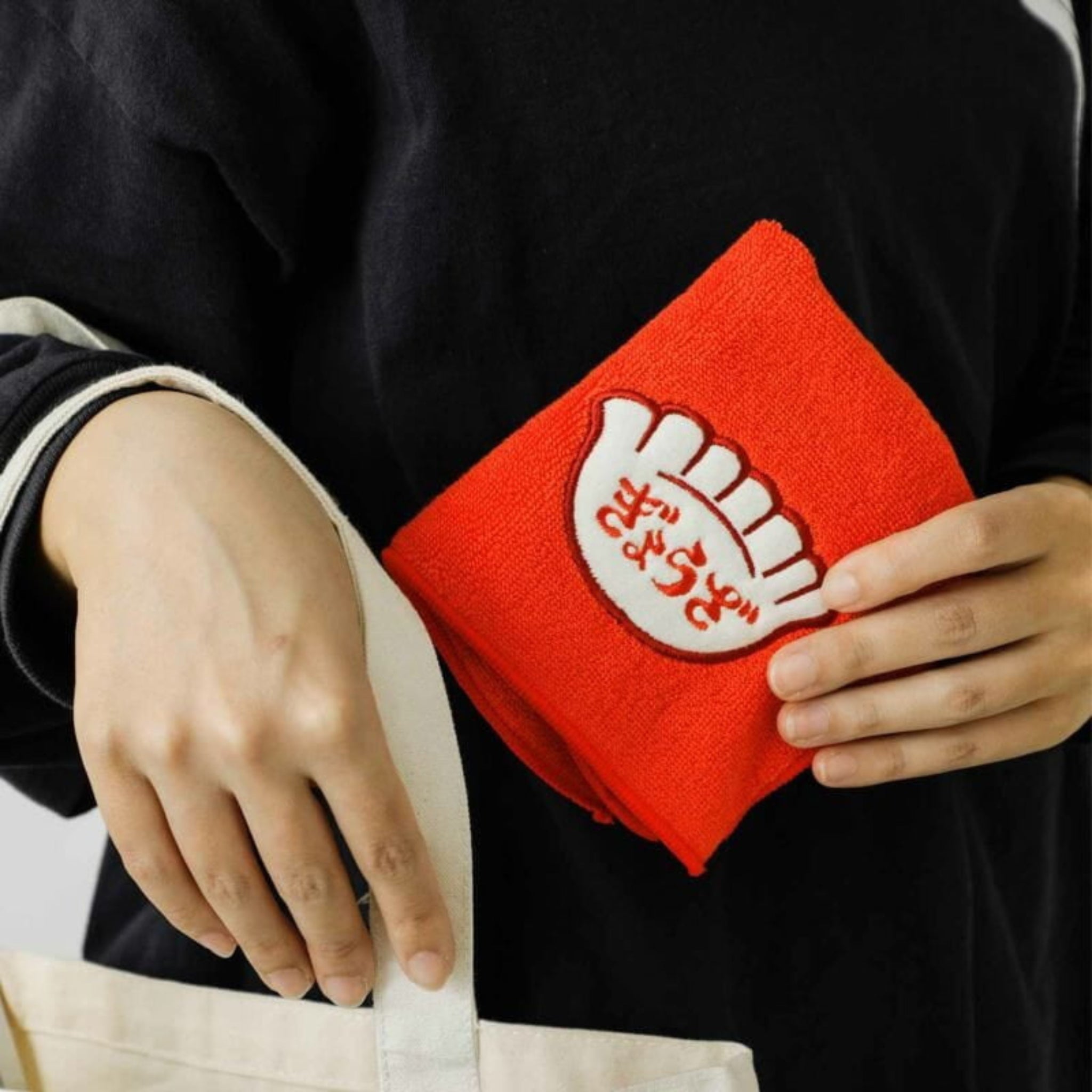 Een persoon plaatst de Hightide New Retro Handdoek Rood 25/25, een rode doek van Hightide met een witte handvormige patch, in een beige tas terwijl hij een donker shirt met lange mouwen en lichtgestreepte manchetten draagt.