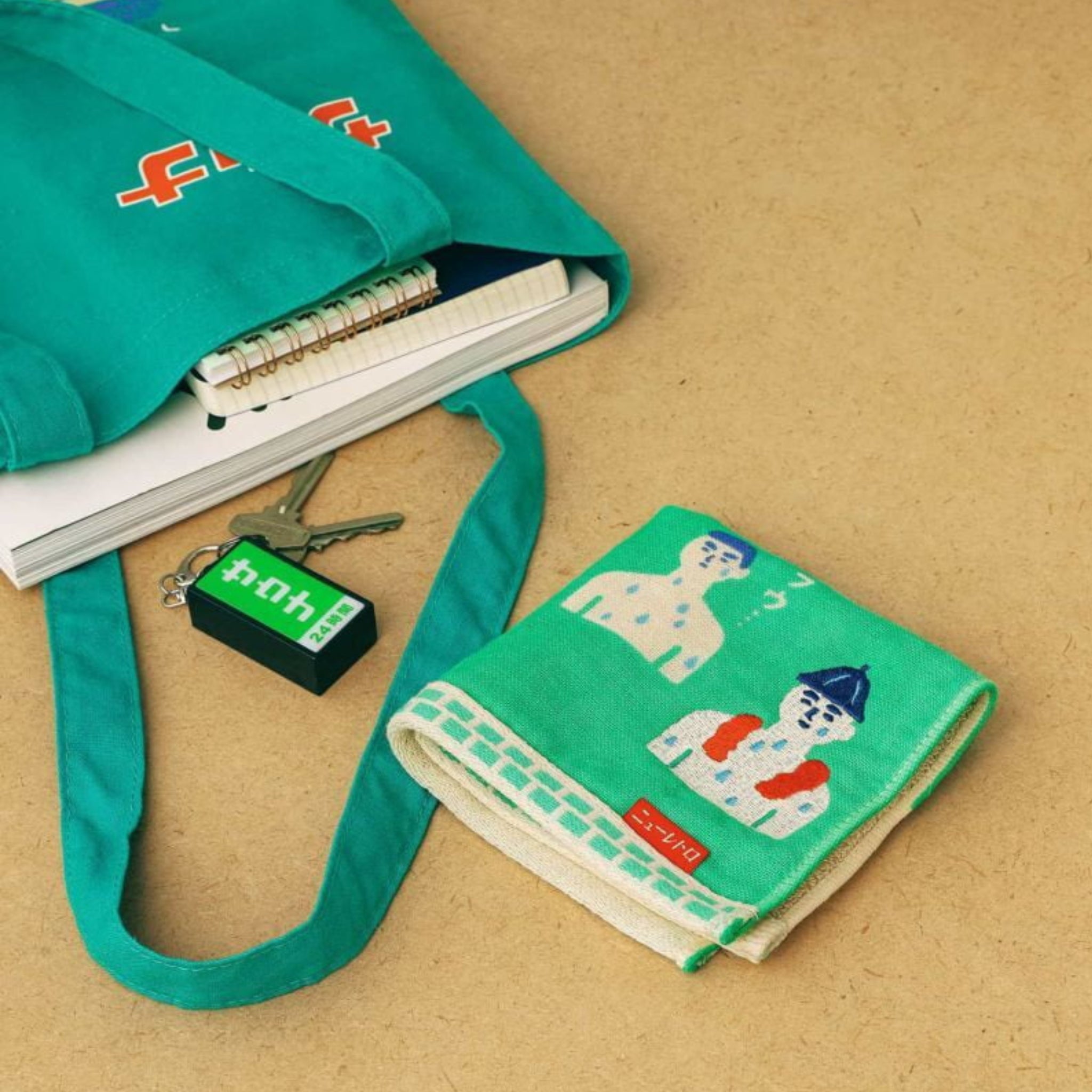 Een groenblauwe draagtas met notitieboekjes, een groene Hightide-sleutelhanger met witte tekst en een gevouwen Hightide Handdoek Sauna 25/25 met geïllustreerde mensen, allemaal afgebeeld op een tan kleurig oppervlak.