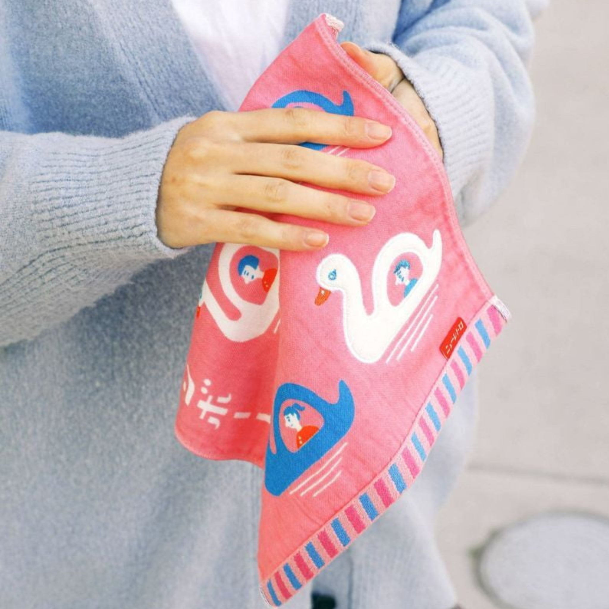 Een persoon in een lichtblauwe trui houdt de Hightide Handdoek Zwaan Boot 25/25 vast, een roze handdoek van Hightide met witte zwanen, kleine figuurtjes en blauw, roze en wit gestreepte randen. De zachte stof maakt het geheel nog aantrekkelijker.