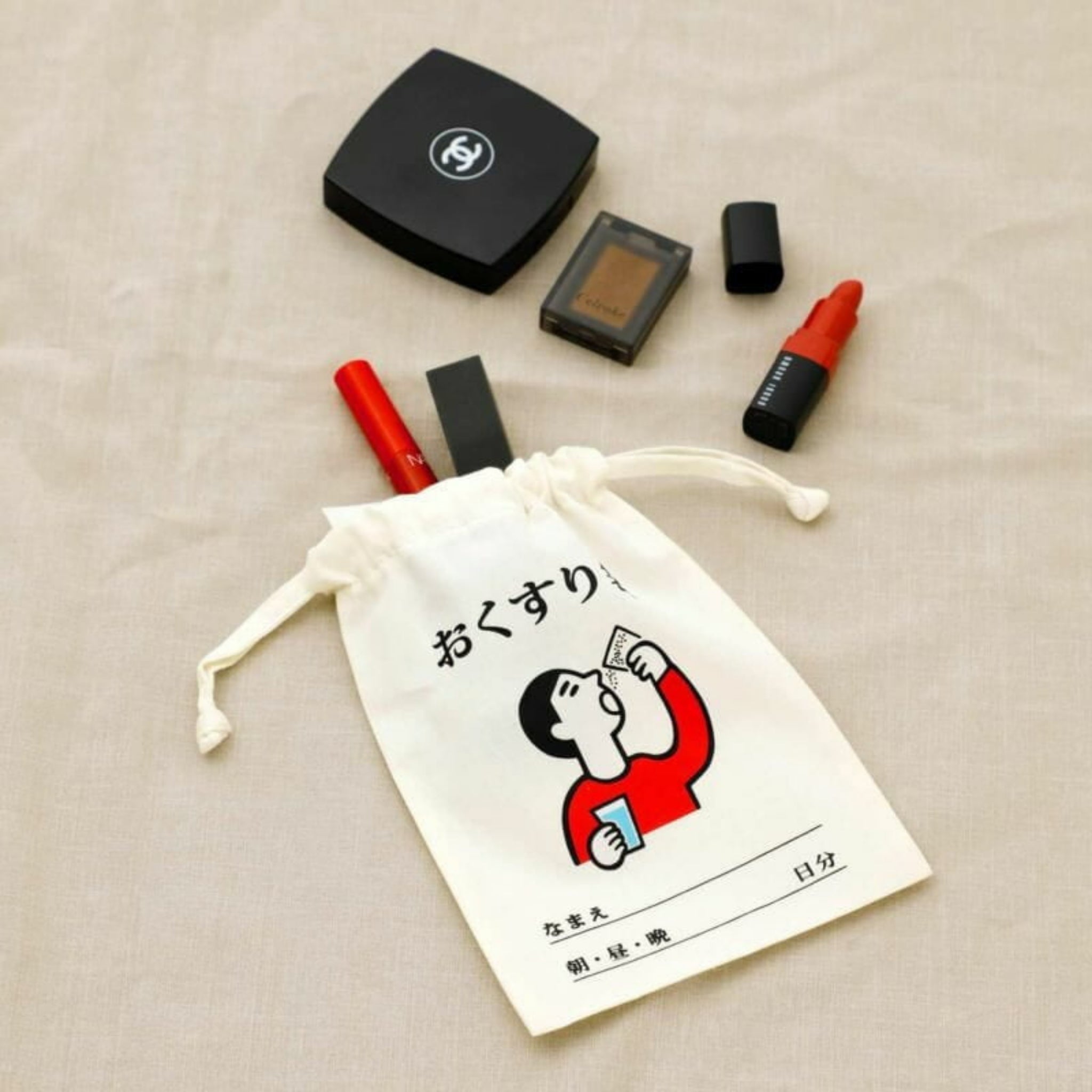 Een Hightide New Retro Katoenen Tas Medicijnen Small van Hightide, met Japanse tekst en een cartoonfiguur die medicijnen neemt, rust op beige stof met een Chanel compact, lippenstift en andere cosmetica in de buurt.