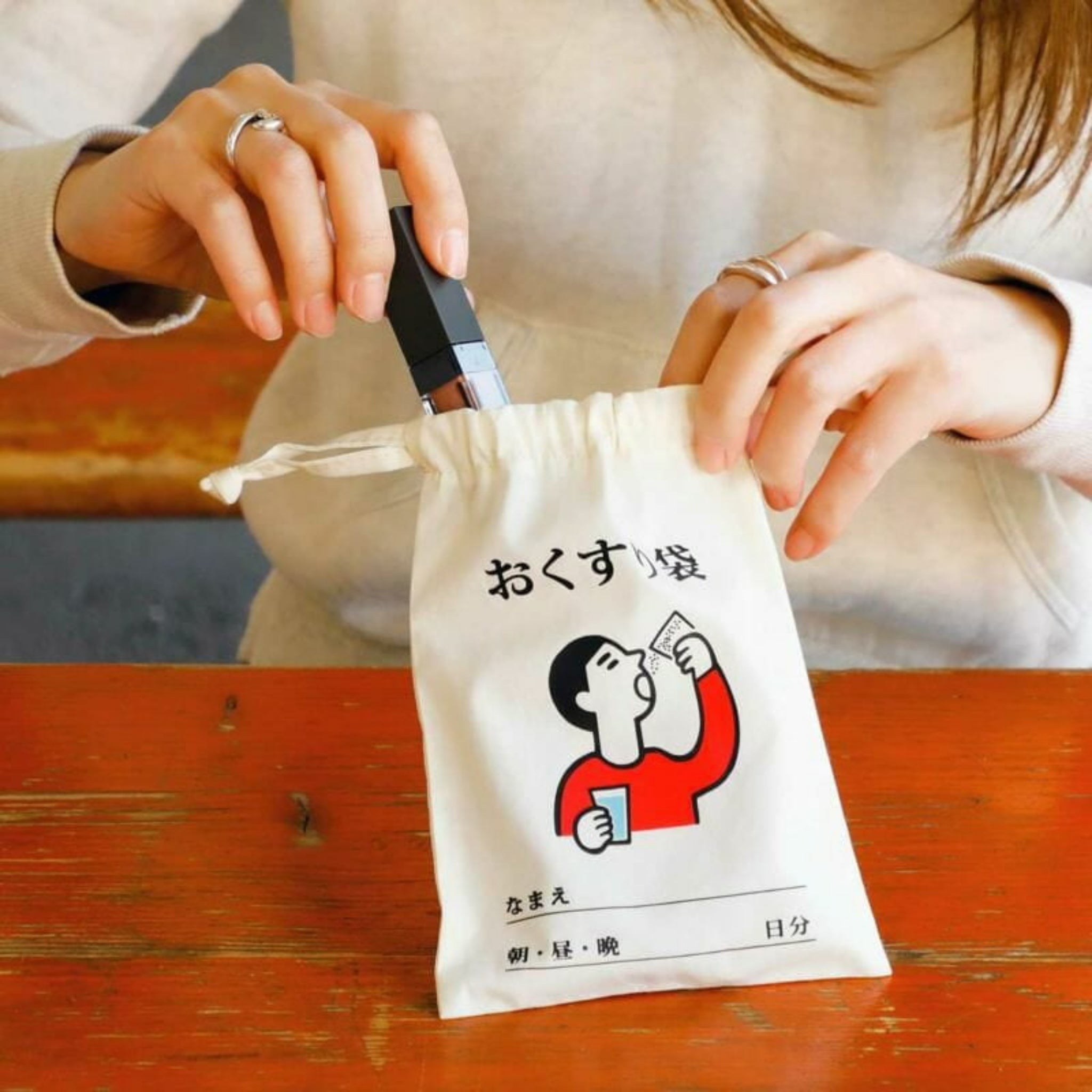 Een persoon houdt de Hightide New Retro Katoenen Tas Medicijnen Small vast, een leuke organizer van Hightide met een cartoon en Japanse tekst, en legt er een voorwerp in om alles netjes op te bergen.