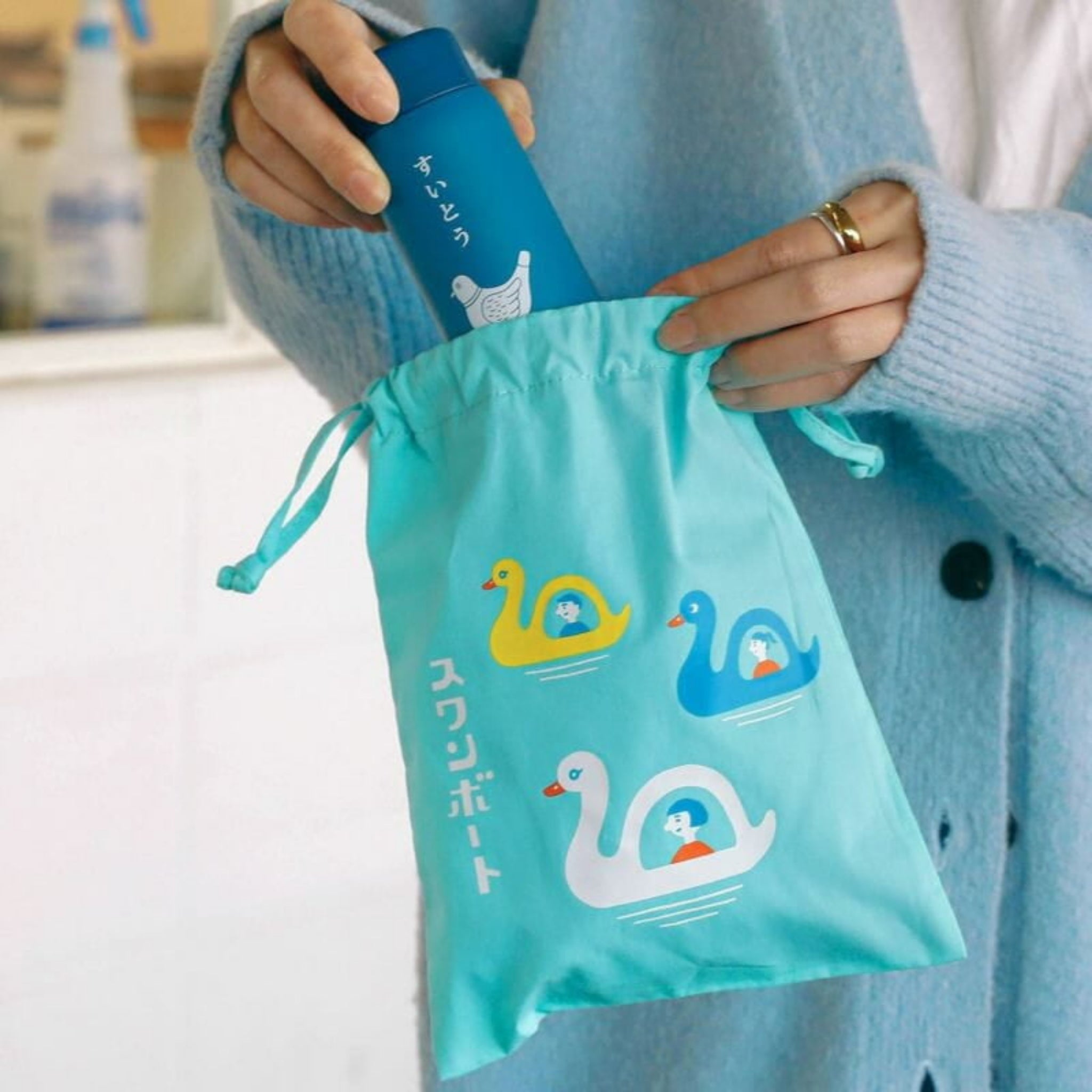 Een persoon in een blauwe jas stopt een blauwe fles met Japanse tekst in een Hightide New Retro Katoenen Tas Zwaan Boot Medium van Hightide, versierd met drie kleurrijke zwanen en Japans schrift.