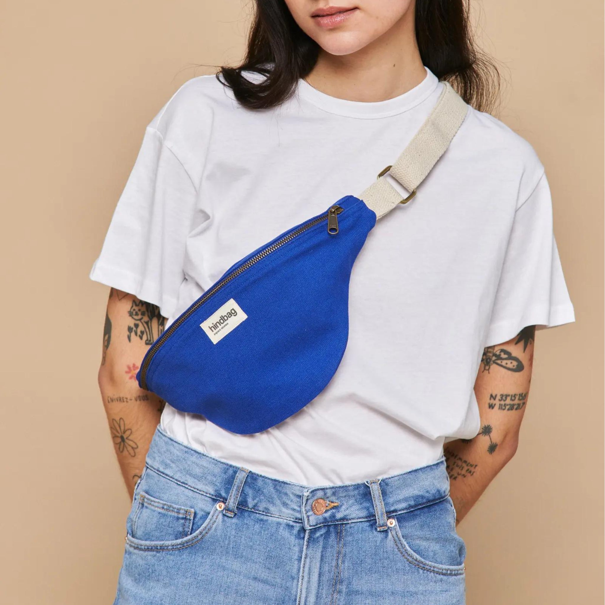 Een persoon in een effen wit t-shirt, blauwe jeans en de Hindbag Olivia Heuptasje Elektrisch Blauw met beige riem van Hindbag staat tegen een beige achtergrond. Op beide armen zijn tatoeages zichtbaar.