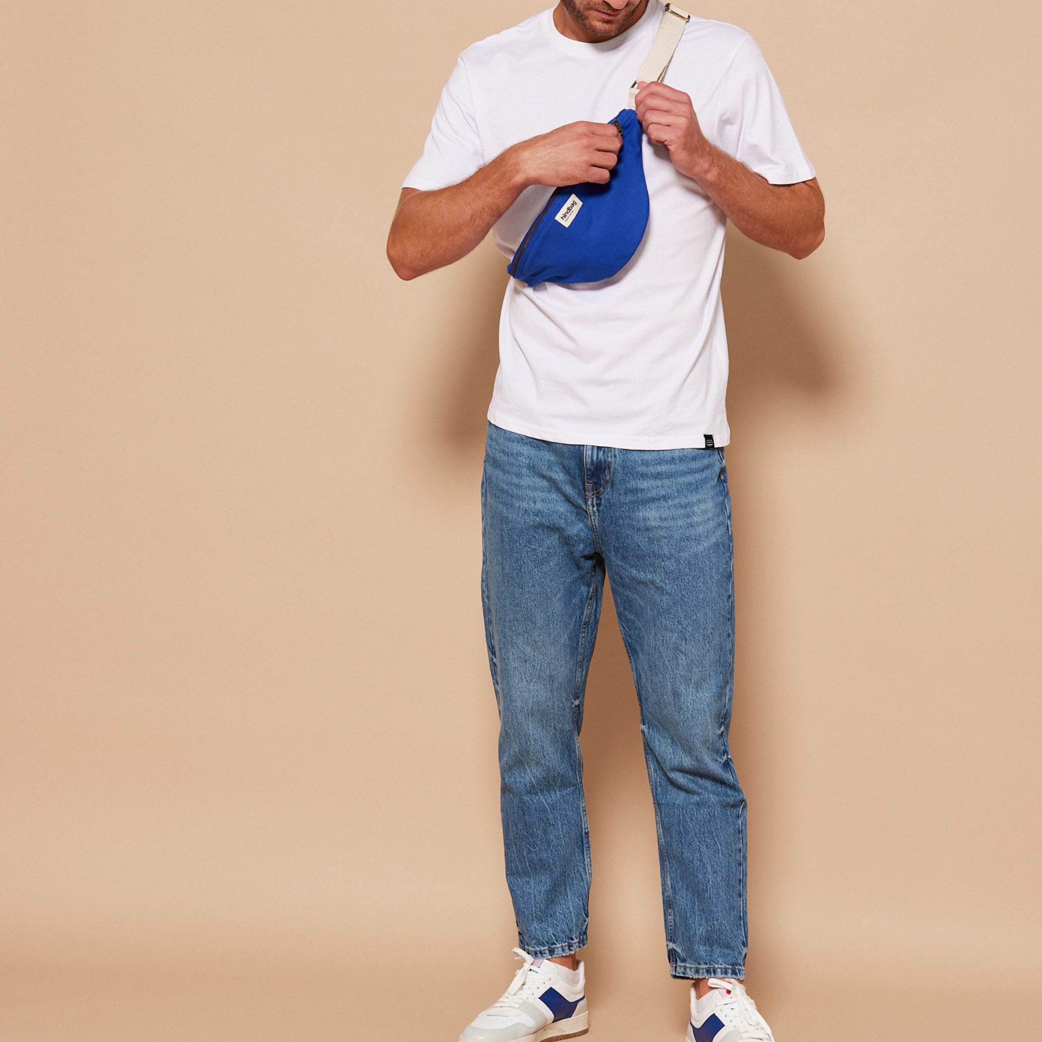 Een man in een wit t-shirt, blauwe jeans en witte sneakers staat tegen een beige achtergrond, terwijl hij een Hindbag Olivia Heuptasje Elektrisch Blauw van Hindbag over zijn borst verstelt. Zijn gezicht is niet zichtbaar.