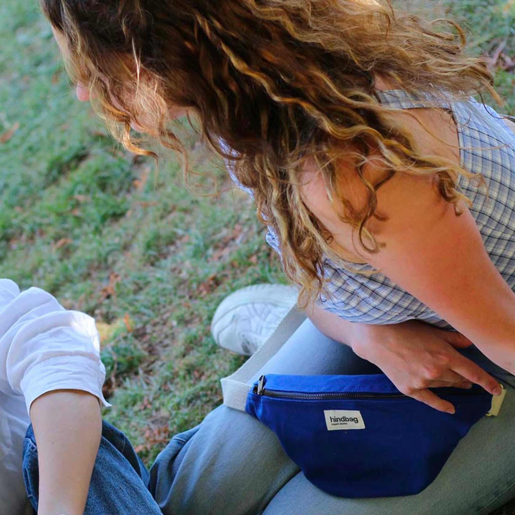 Een persoon met krullend haar in een blauw geruite top zit op gras en houdt een Hindbag Olivia Heuptasje Elektrisch Blauw van Hindbag vast. Iemand anders in lichte kleding is gedeeltelijk zichtbaar naast hen.