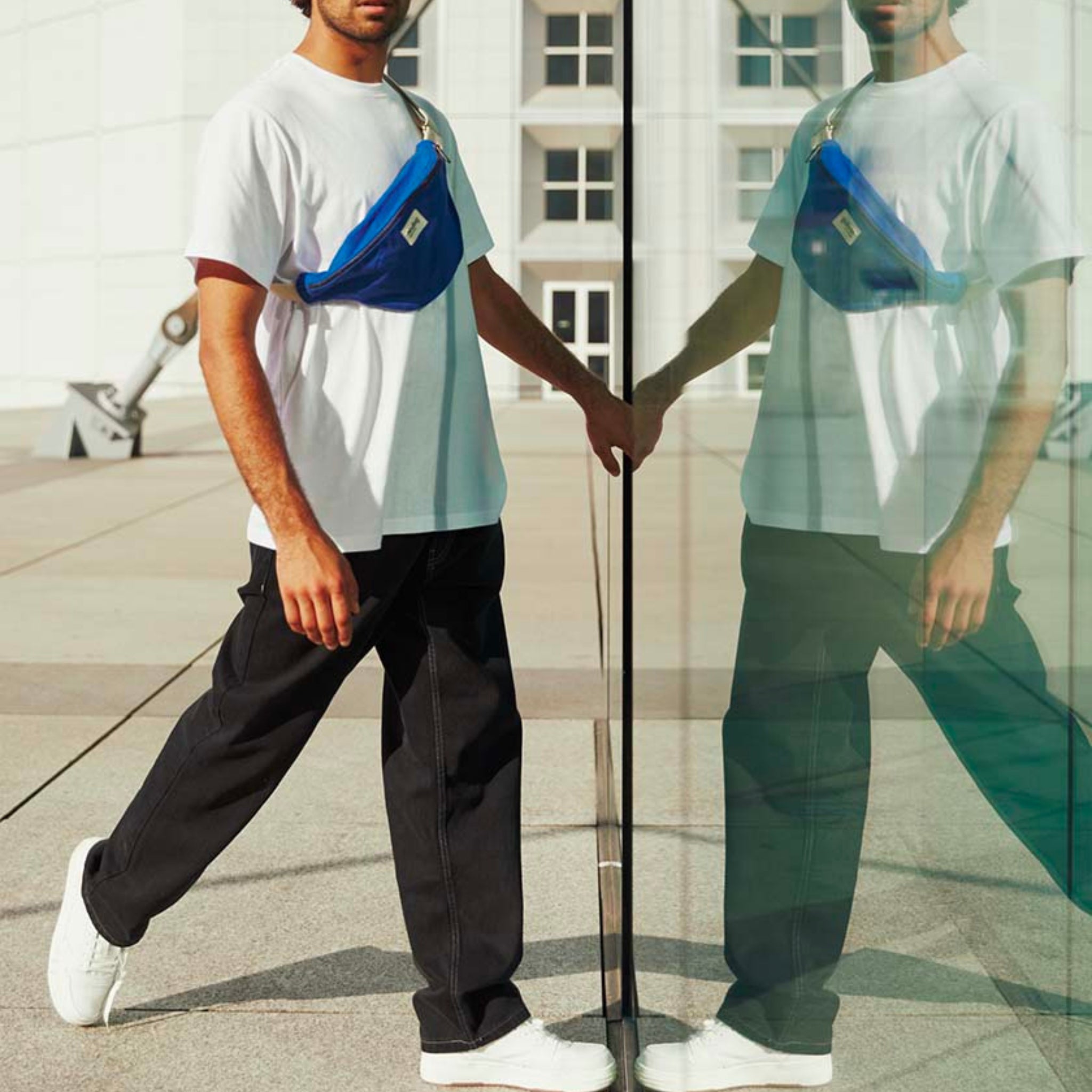 Een man in een wit t-shirt, zwarte broek, witte sneakers en een Hindbag Olivia Heuptasje Elektrisch Blauw loopt terwijl hij een glazen wand aanraakt, waardoor een duidelijke reflectie van zichzelf ontstaat.