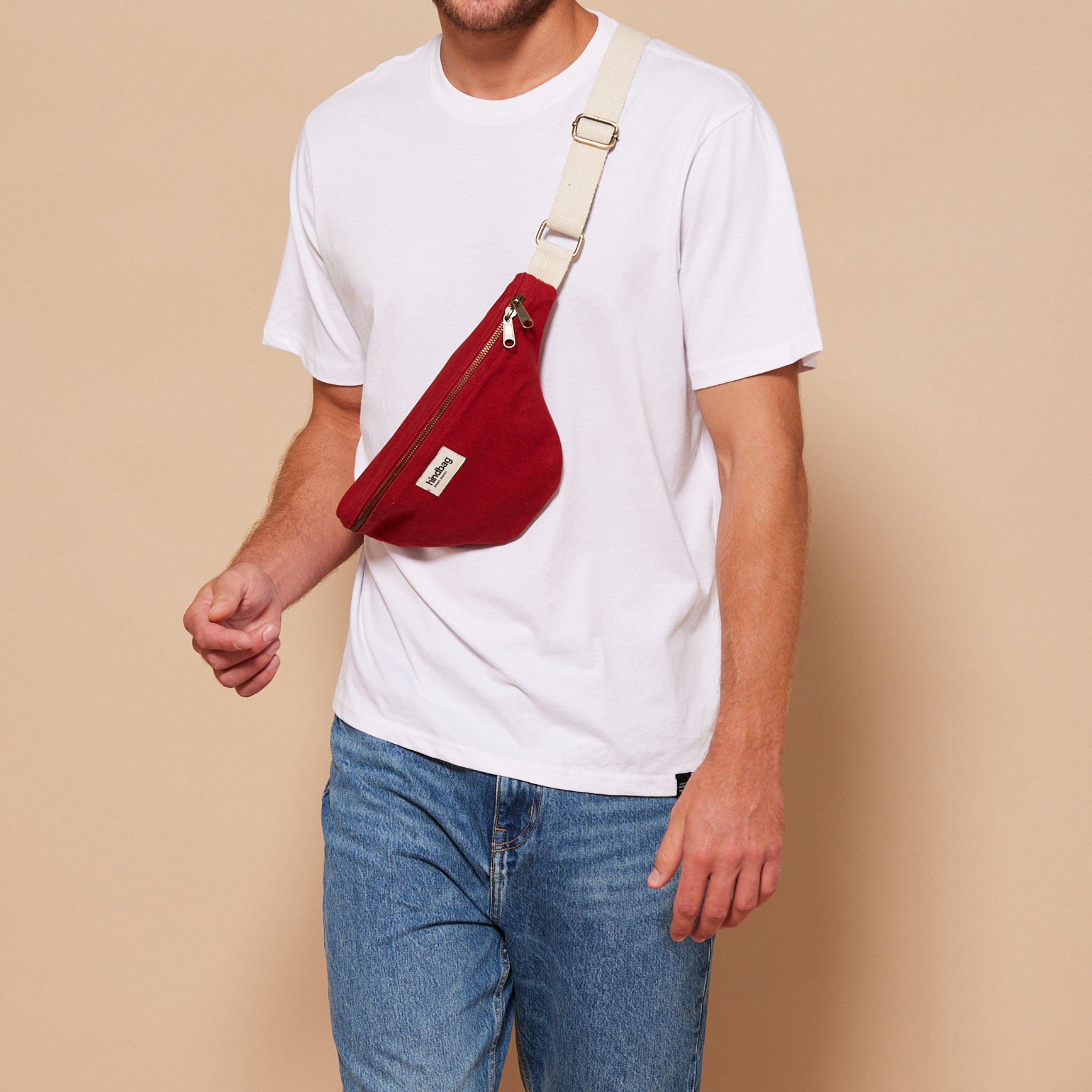 Een persoon in een effen wit T-shirt en blauwe jeans draagt de Hindbag Olivia Heuptasje Terracotta, gemaakt van biologisch katoen, met een beige riem over de borst, staande tegen een effen beige achtergrond.