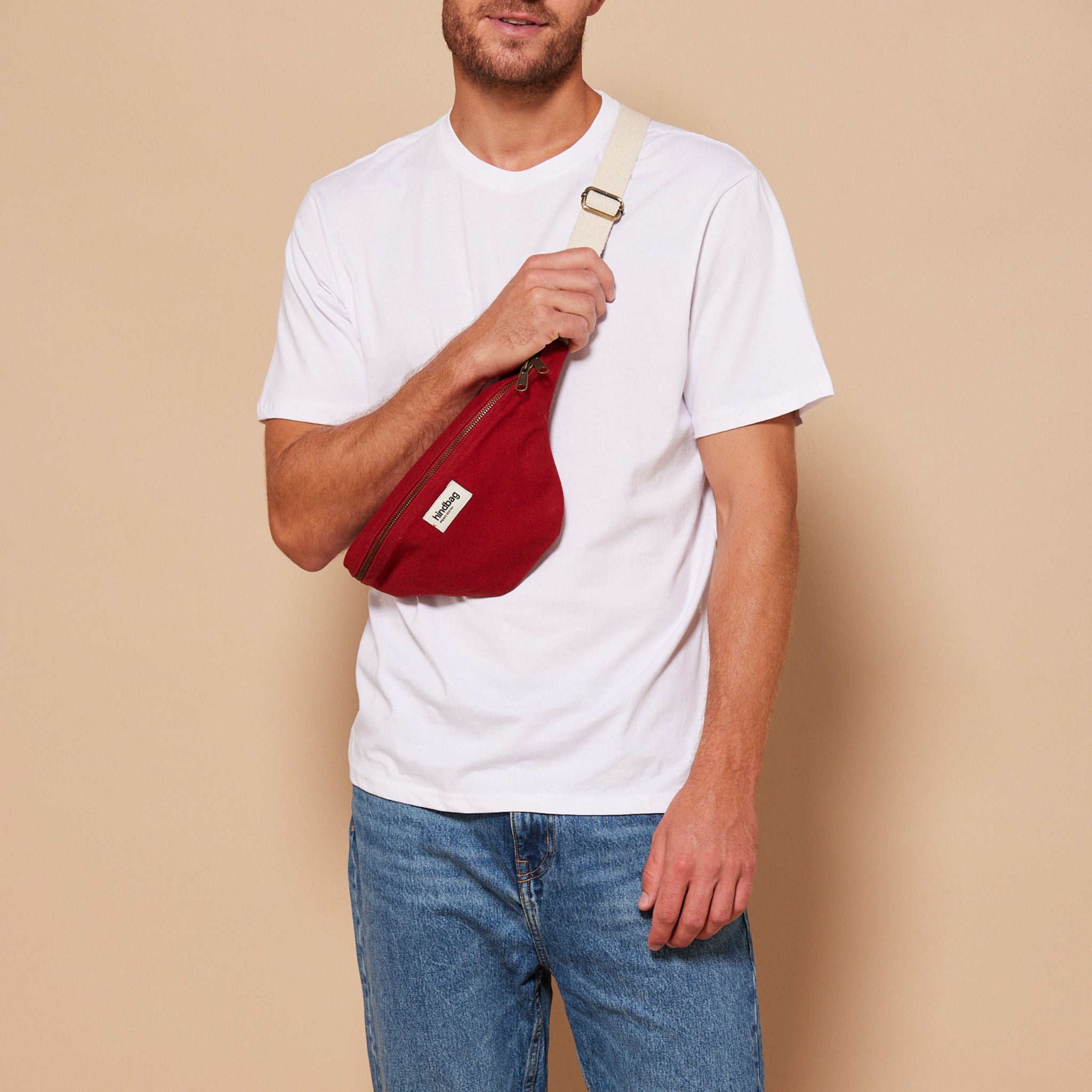 Een man in een effen wit t-shirt en een blauwe spijkerbroek staat tegen een beige achtergrond. Hij draagt de Hindbag Olivia Heuptasje Terracotta van Hindbag met een crèmekleurige riem over zijn borst. De tas is gemaakt van organisch katoen. Zijn gezicht is gedeeltelijk buiten beeld.