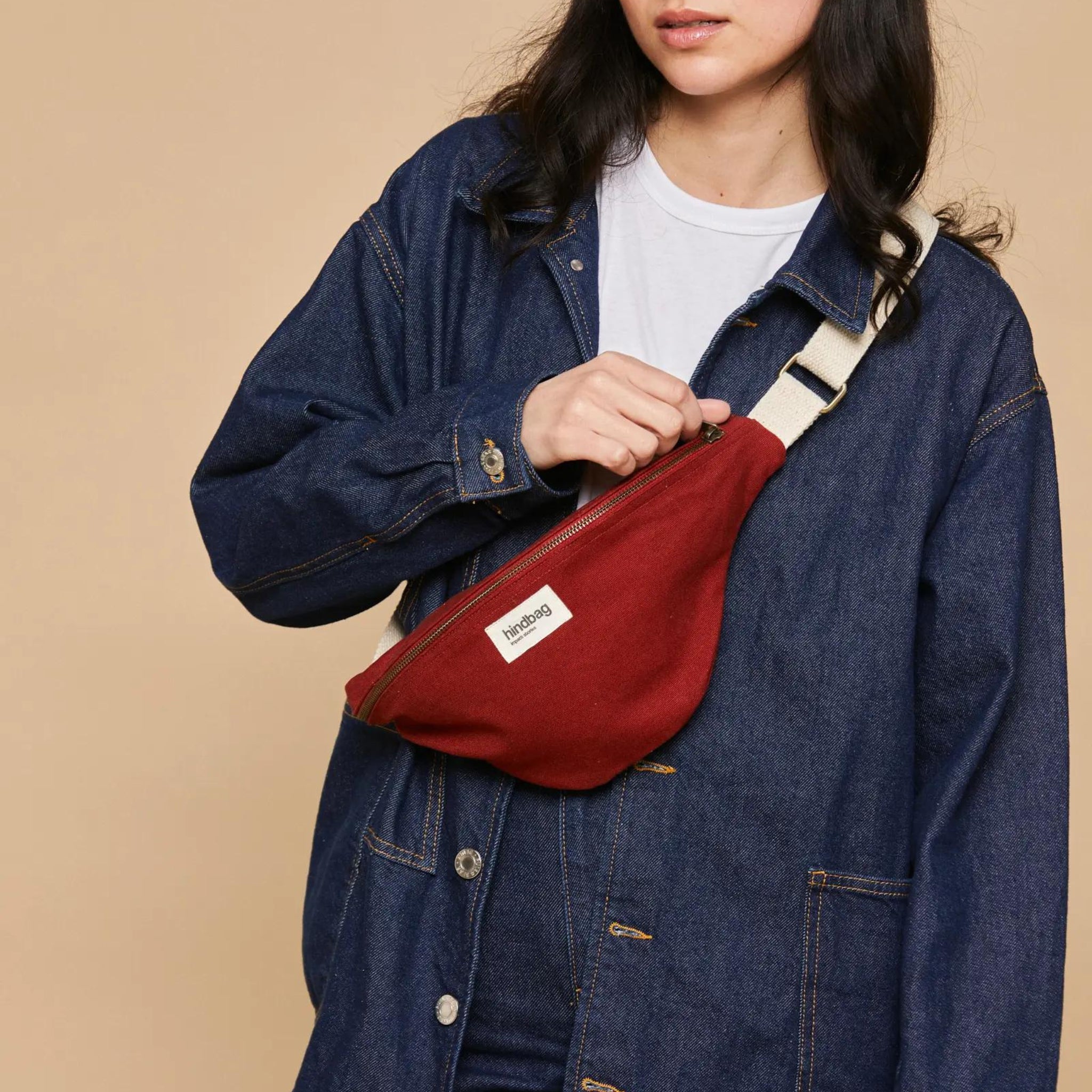 Een persoon in een donker spijkerjasje over een wit shirt houdt de riem vast van de Hindbag Olivia Heuptasje Terracotta, gemaakt van biologisch katoen met een witte rits en logolabel, staande tegen een beige achtergrond.