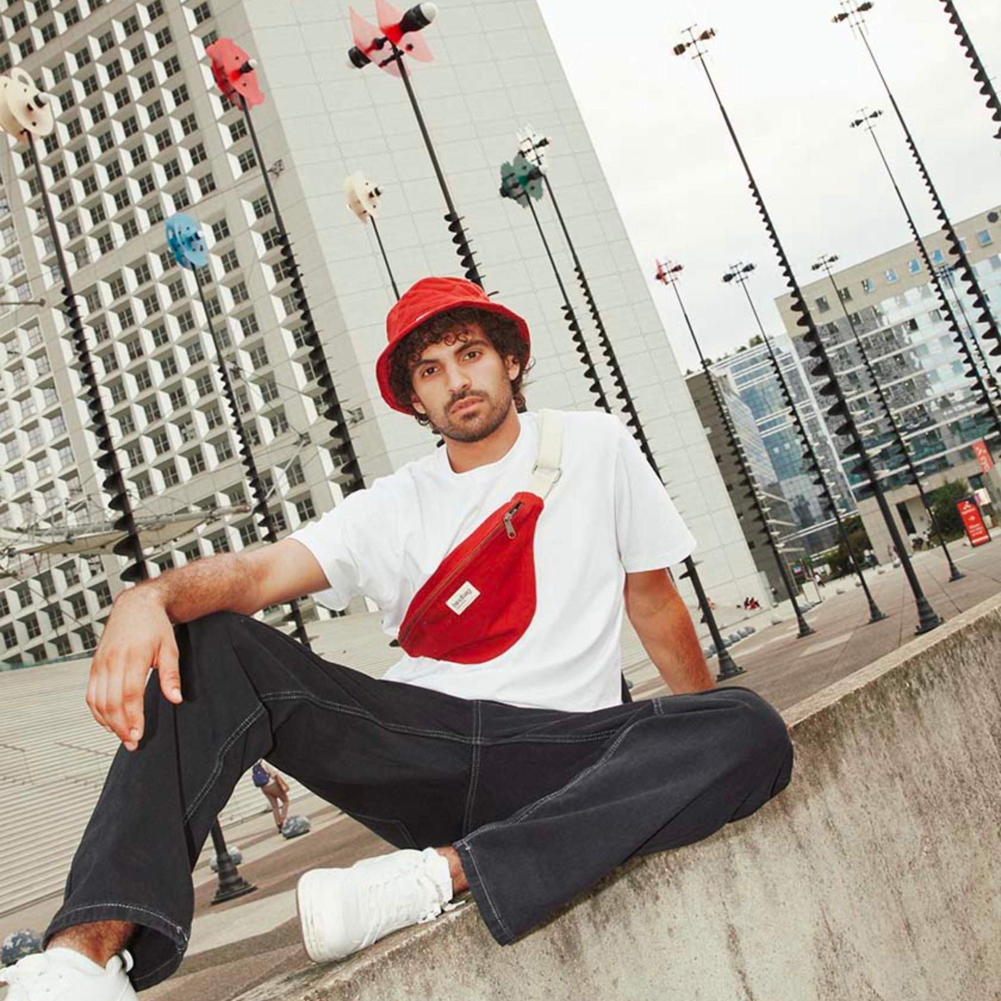 Een jongeman met een rode emmerhoed, een wit T-shirt van organisch katoen, een zwarte broek, witte sneakers en de Hindbag Olivia Heuptasje Terracotta zit op een laag betonnen muurtje in een stedelijk gebied met moderne gebouwen en straatverlichting.