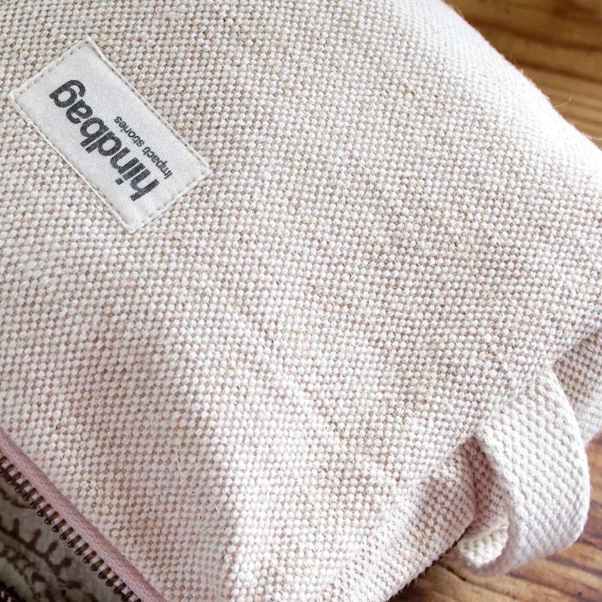 Een close-up van de Hindbag Leon Toilettas Jute van Hindbag, gemaakt van lichtbeige biologisch katoen met een zichtbare rits en merklabel, rustend op een houten oppervlak.