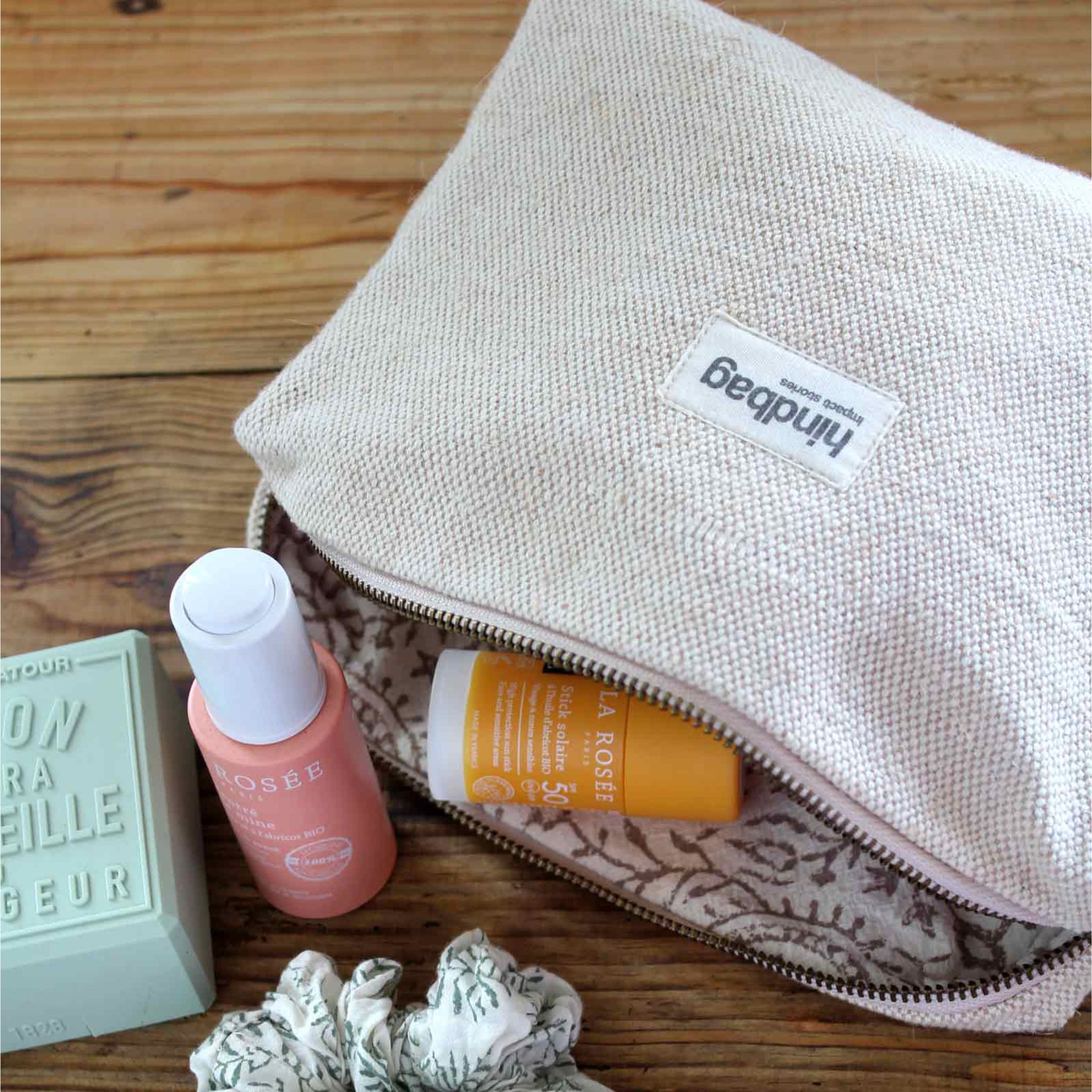 Een beige Hindbag Leon Toilettas Jute van Hindbag van jute ligt open op een houten oppervlak. Ernaast liggen een gele tube, een roze spuitfles, een groen stuk zeep en een gebloemde scrunchie.